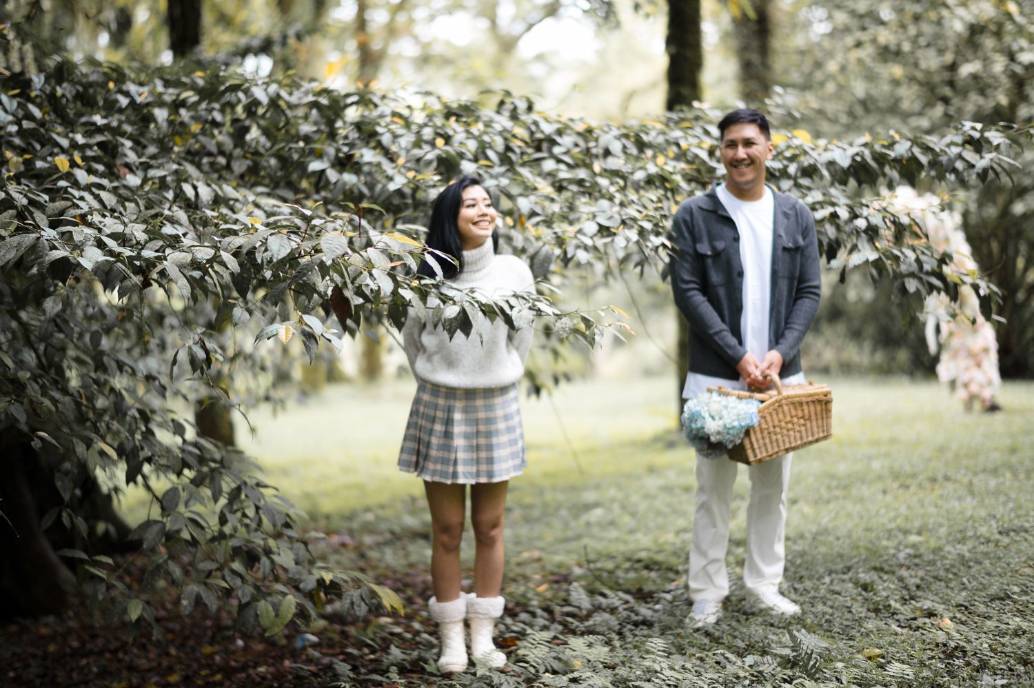 Couple enjoying a picnic in a lush green park, woman holding leaves, man carrying a wicker basket.