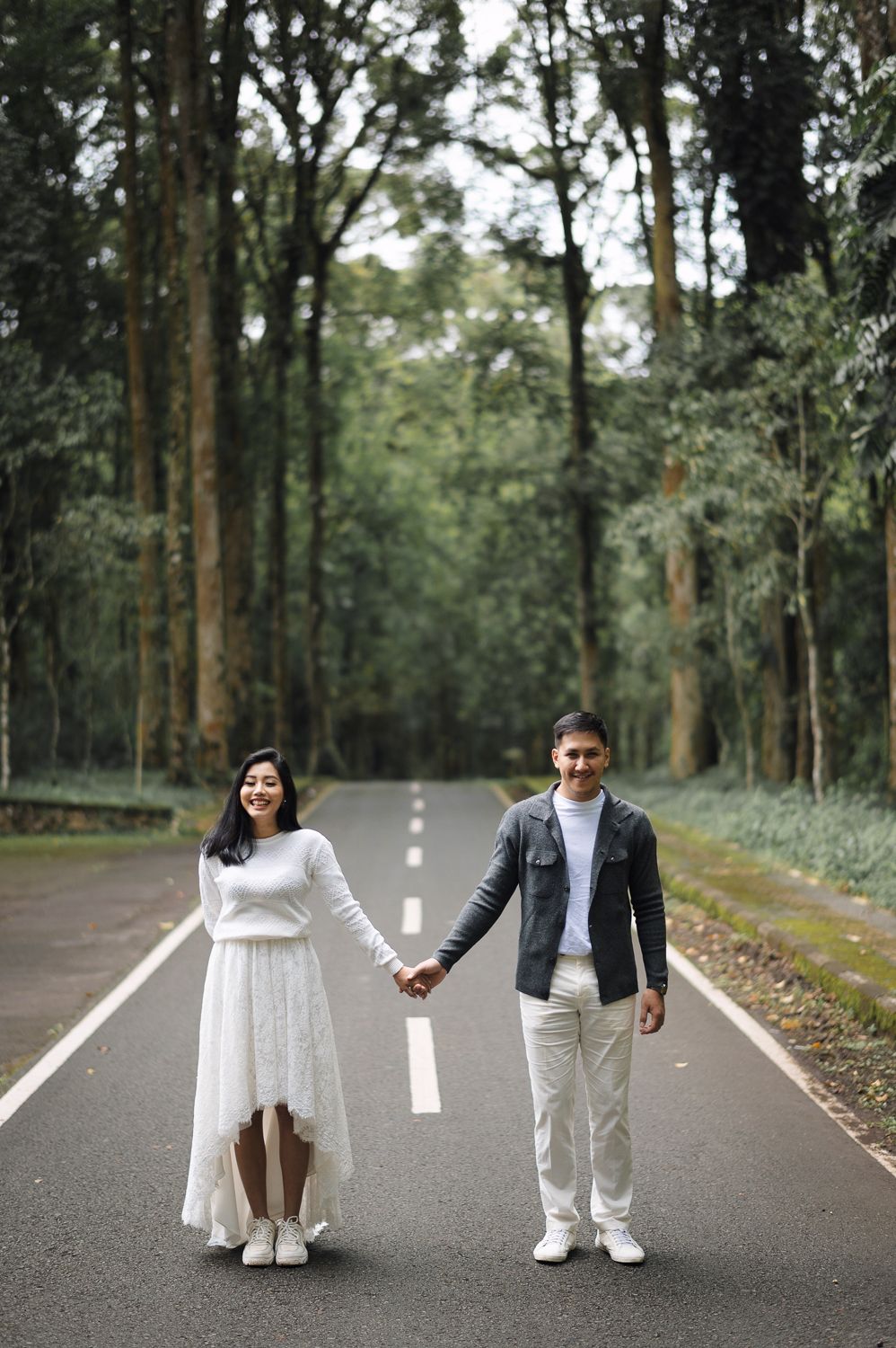 Couple holding hands on a tree-lined road, woman in white dress and man in casual attire, outdoor pre-wedding photo.