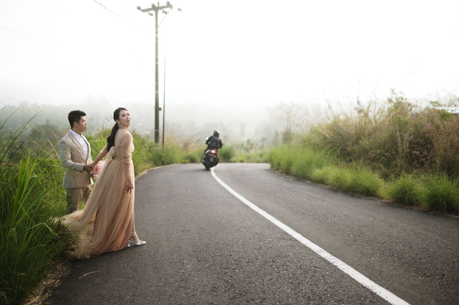 Couple in wedding attire posing on a misty road with a motorcyclist in the background.