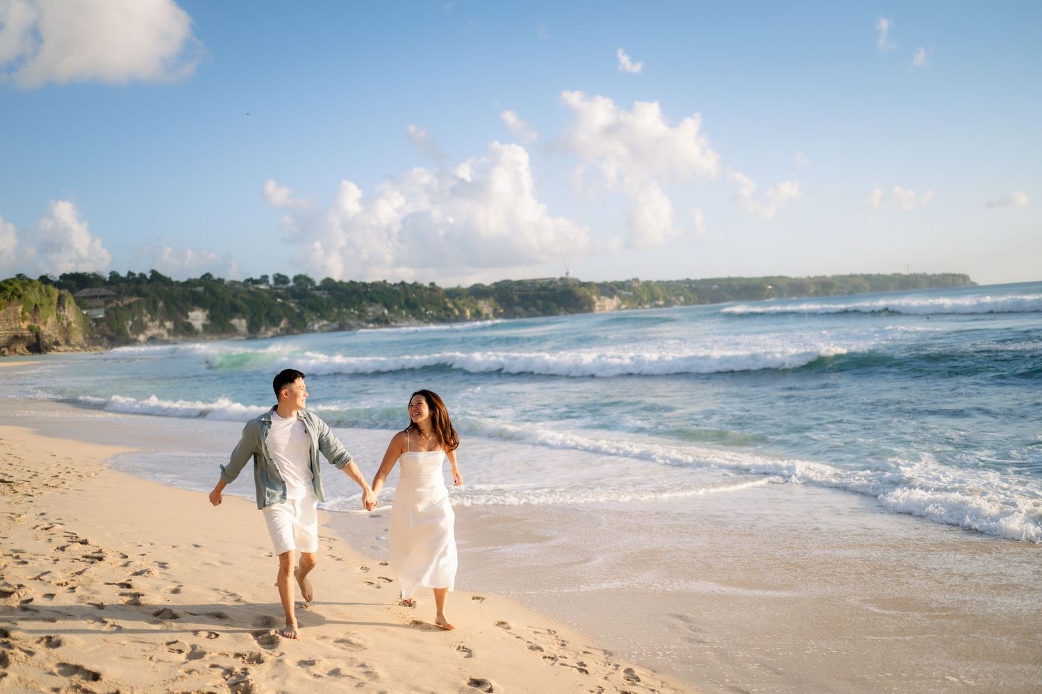 A couple holds hands and walks along a sunny tropical beach with turquoise waves and lush cliffs in the background.
