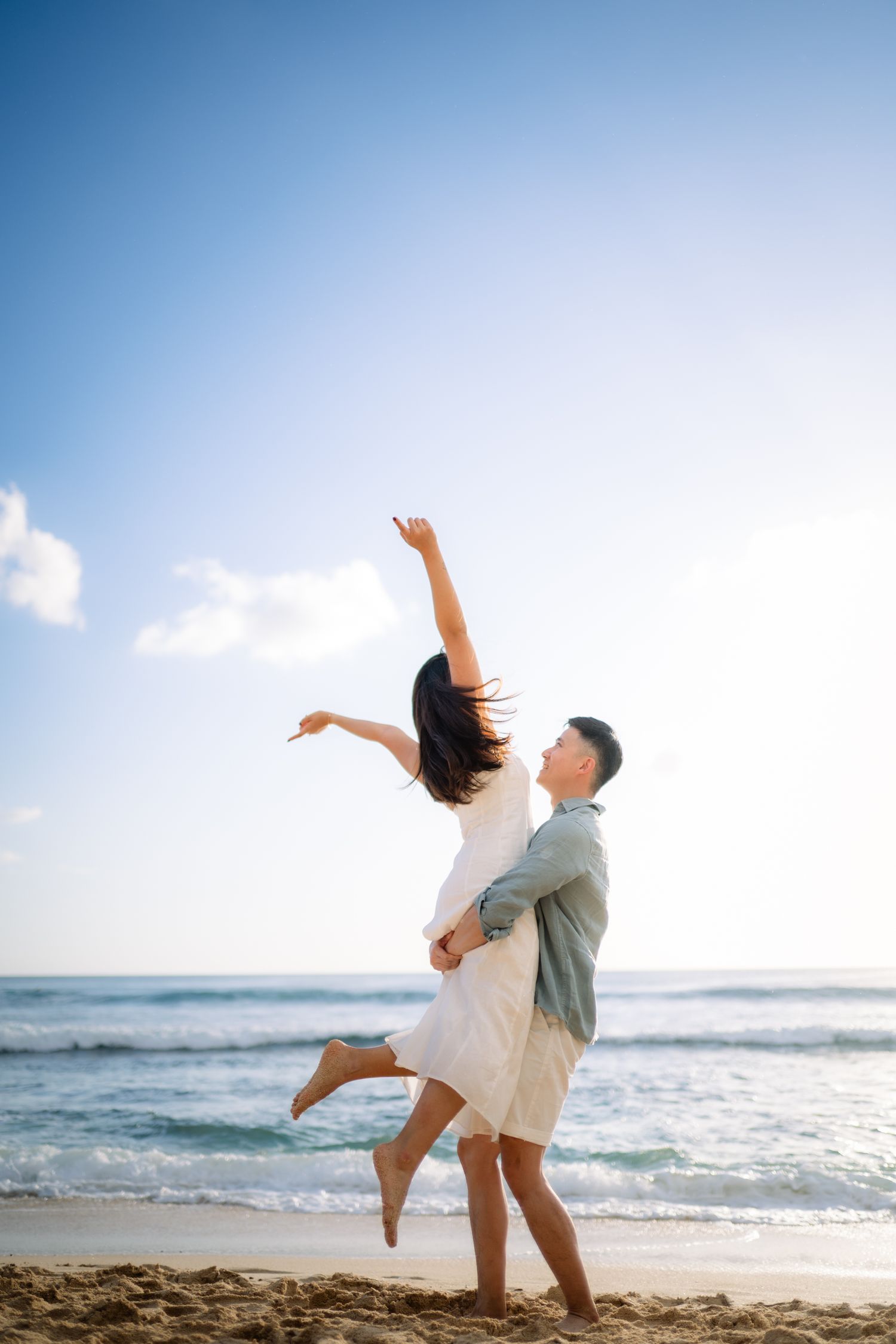 Couple dancing joyfully on a sunny beach, man lifting woman in white dress with ocean waves in the background.