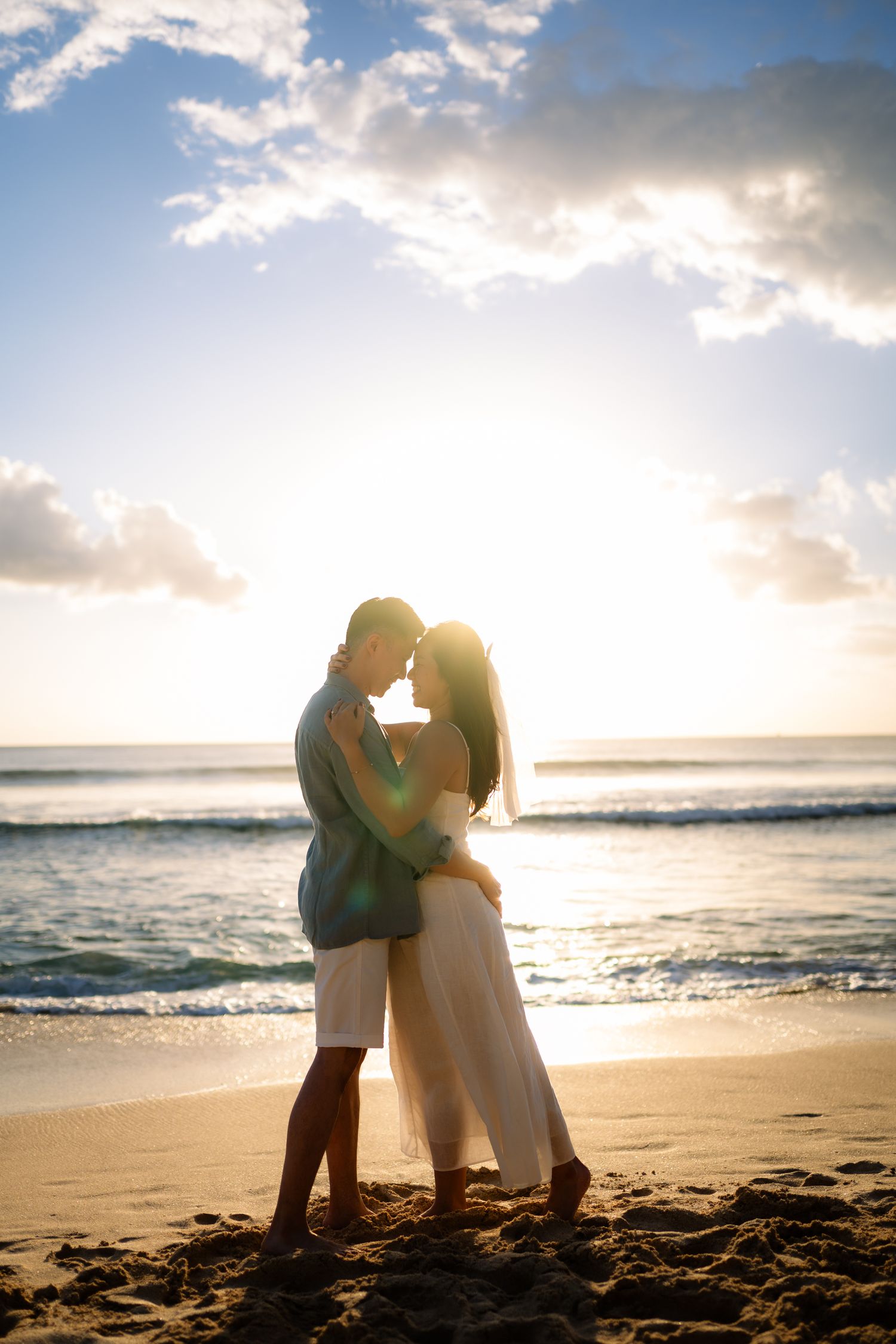 Couple embracing on a sandy beach at sunset with ocean waves and dramatic cloudy sky backdrop.