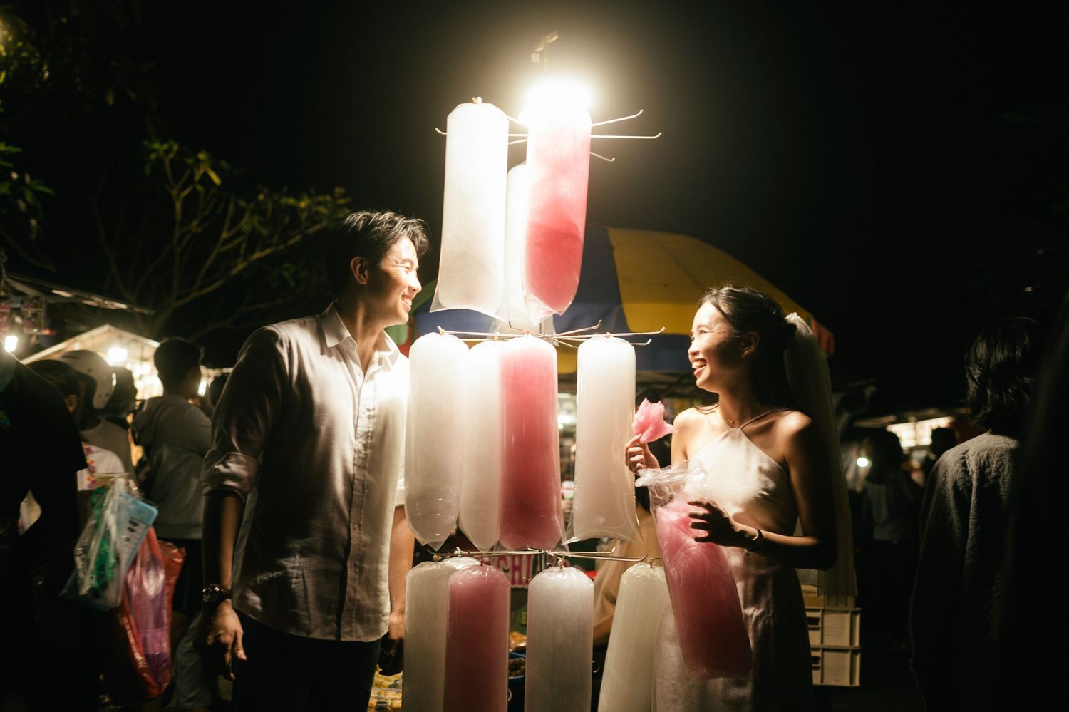 Couple smiling at a colorful illuminated popsicle stand at a night market.