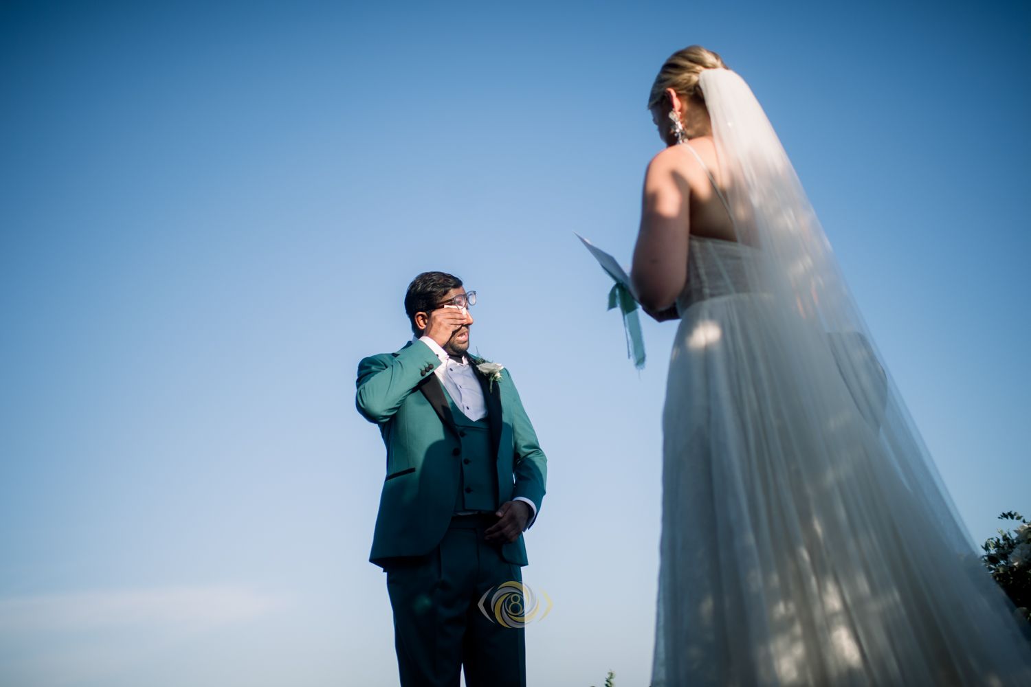 Emotional groom in teal suit wiping tears seeing bride in white wedding dress and veil against clear blue sky.