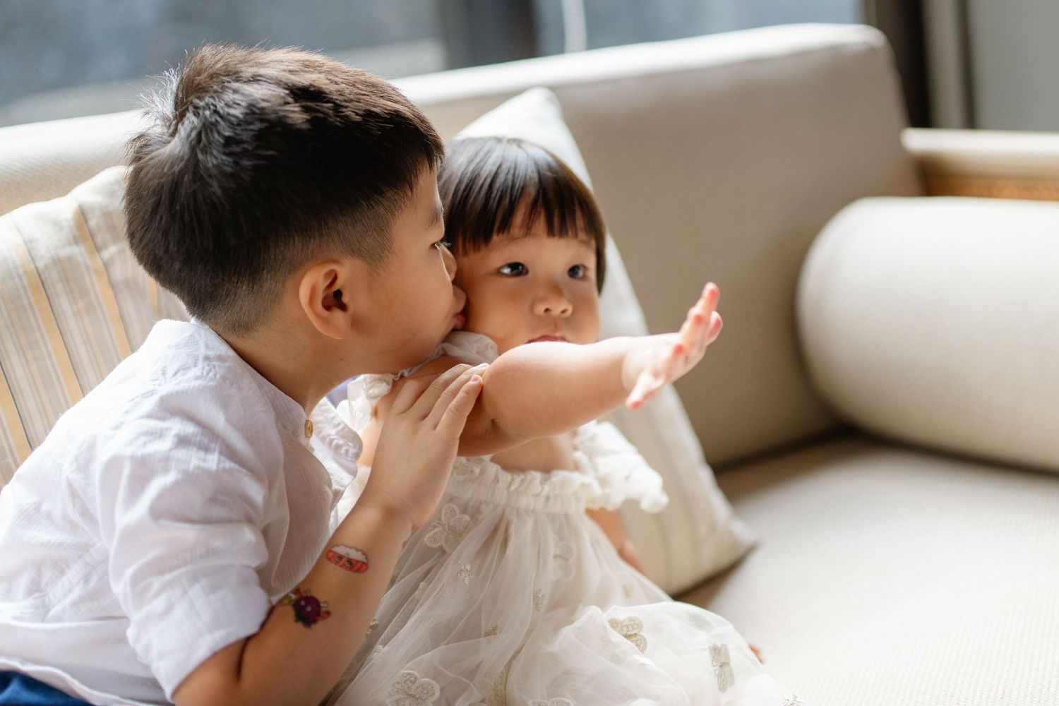 A young boy kisses a toddler girl on the cheek as she sits on a beige sofa in white outfits.