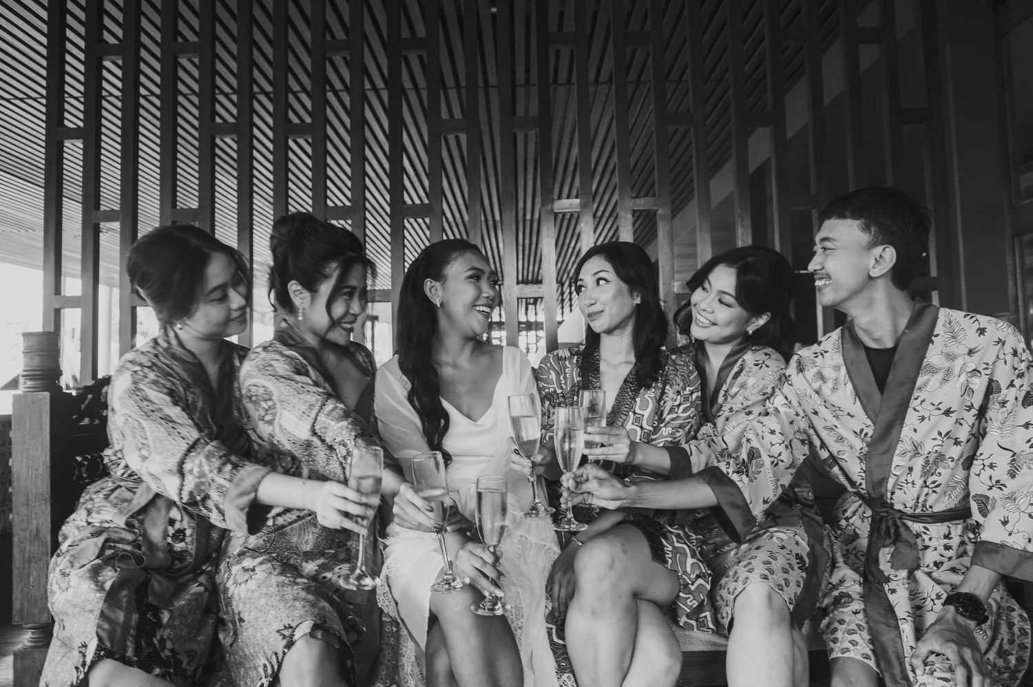 Bridesmaids in floral robes toasting with champagne glasses, laughing together in black and white photo.