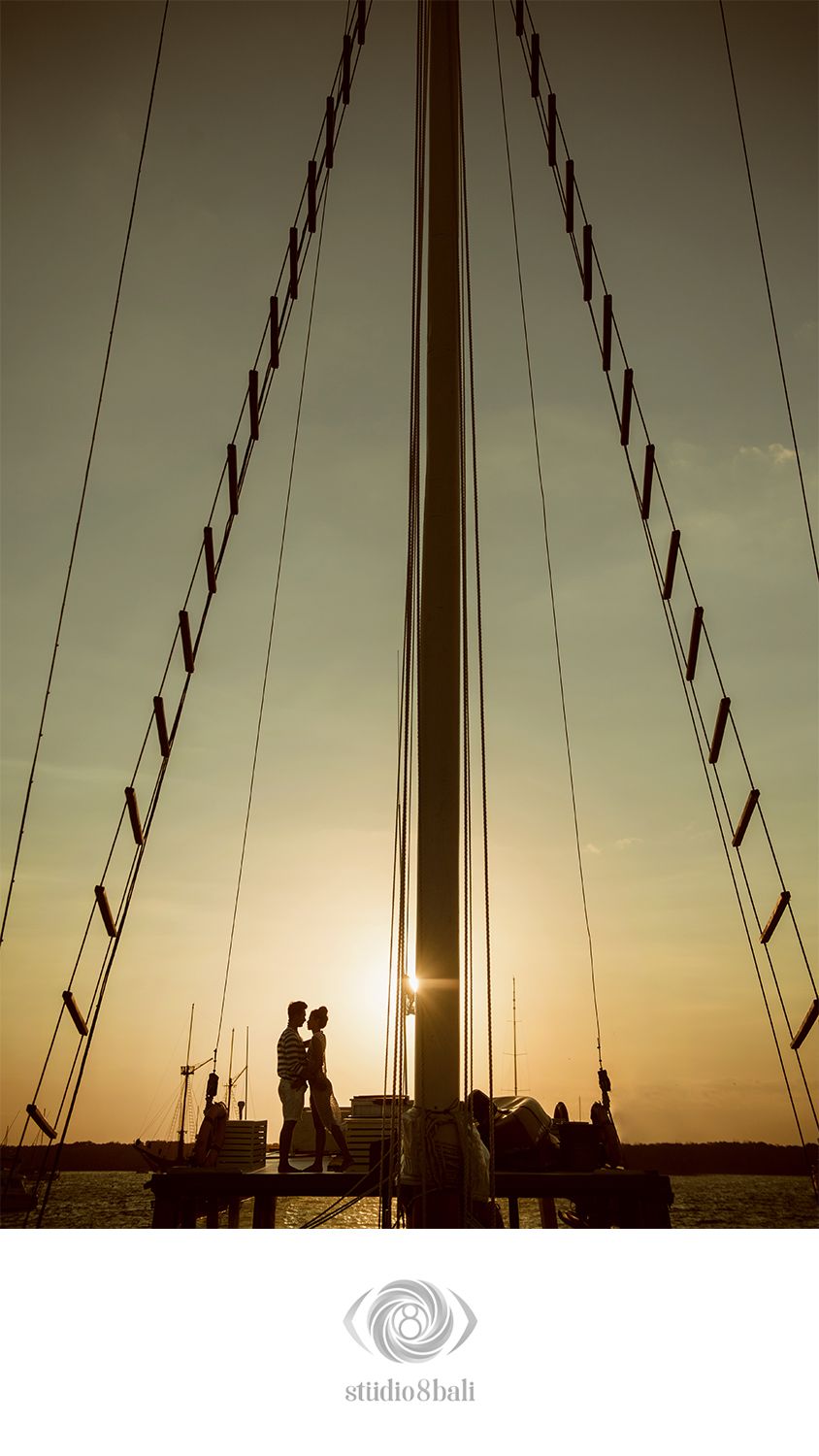 Couple silhouetted against golden sunset beneath sailboat mast and rigging on water.