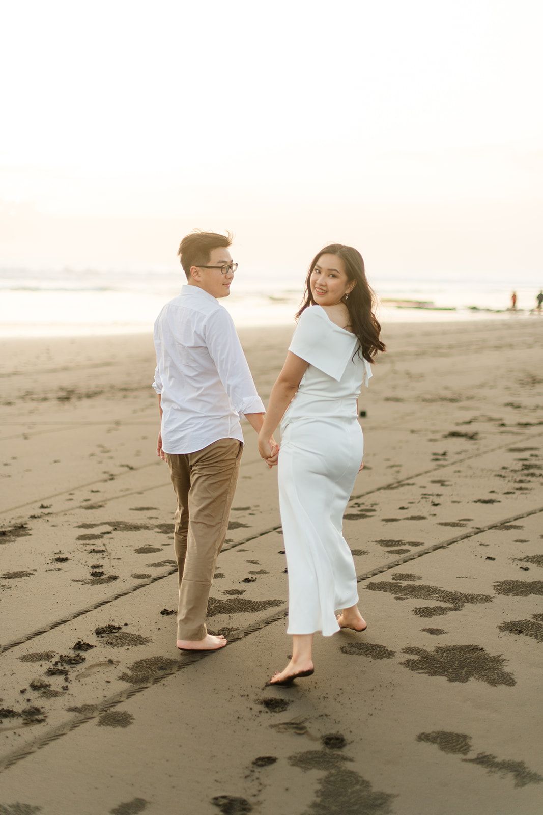 Couple holding hands walking on beach at sunset, woman in white dress looking back, man in white shirt and khaki pants.