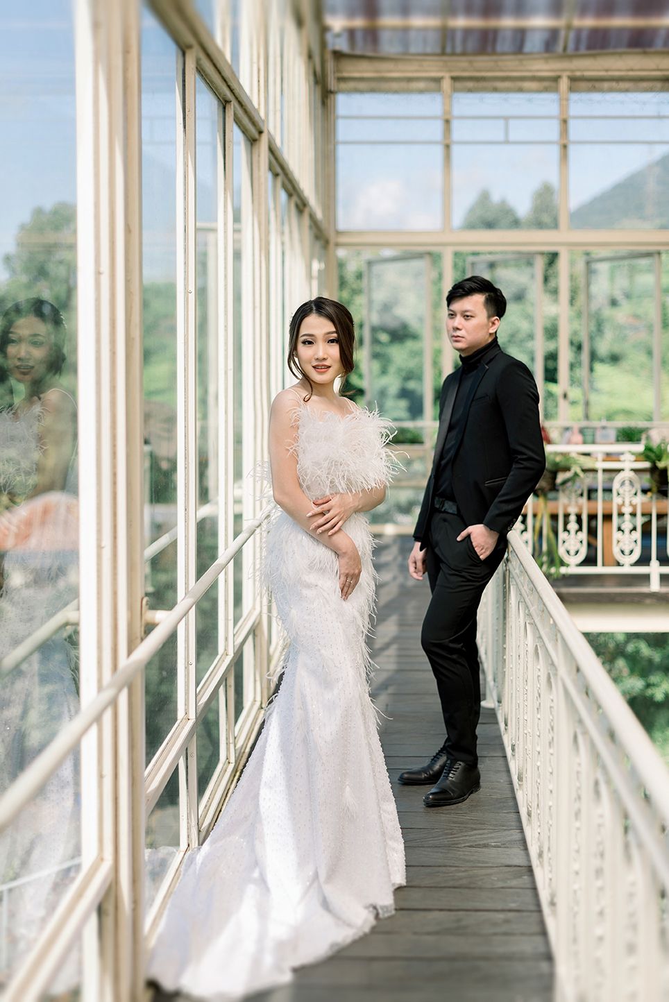 A bride in a white feathered gown and groom in black suit pose on a sunlit glass corridor with green hills backdrop.