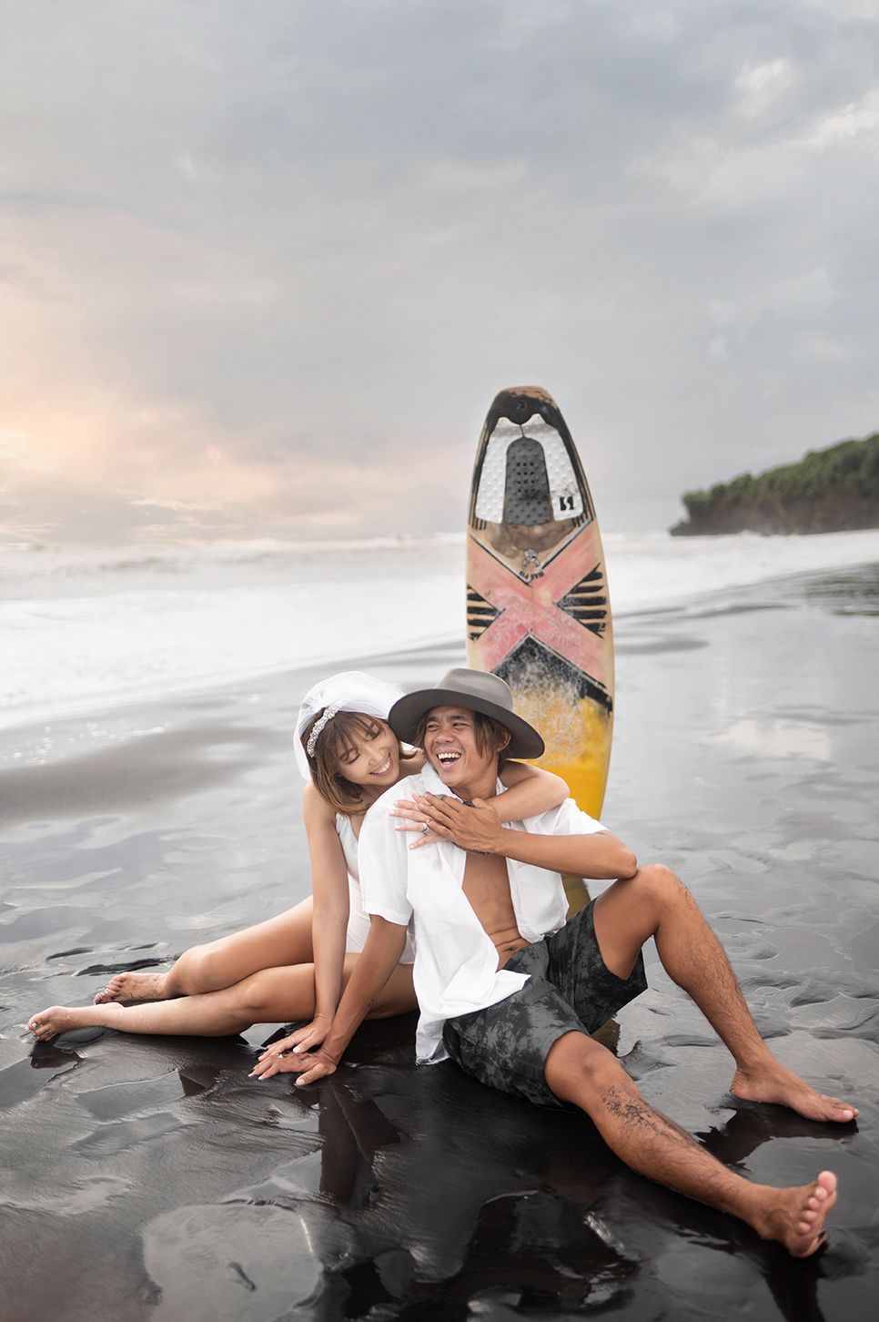 A couple sits laughing on a black sand beach with a colorful surfboard, sharing a joyful romantic moment by the ocean.