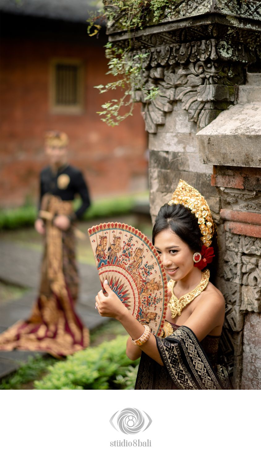 Balinese bride in traditional attire holds decorative fan near stone temple wall, groom stands in background.
