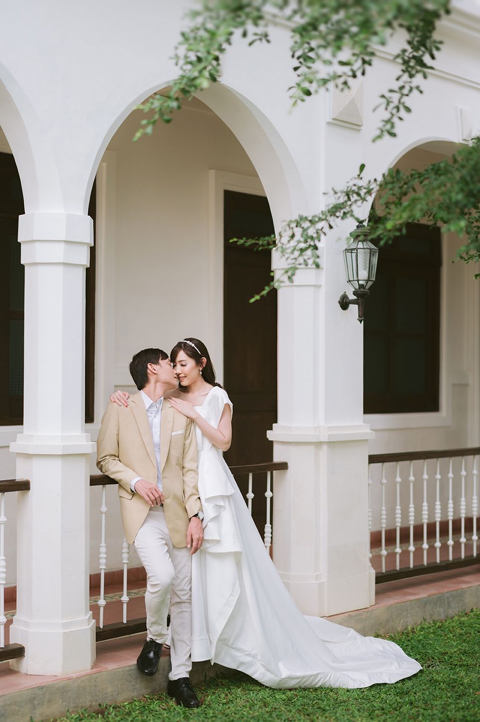 Bride in white gown and groom in beige suit share a romantic kiss on elegant colonial building veranda.
