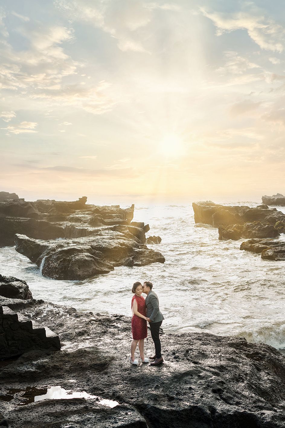 Couple kissing on rocky shoreline with crashing waves and stunning sunburst sky in a vibrant outdoor engagement photo.