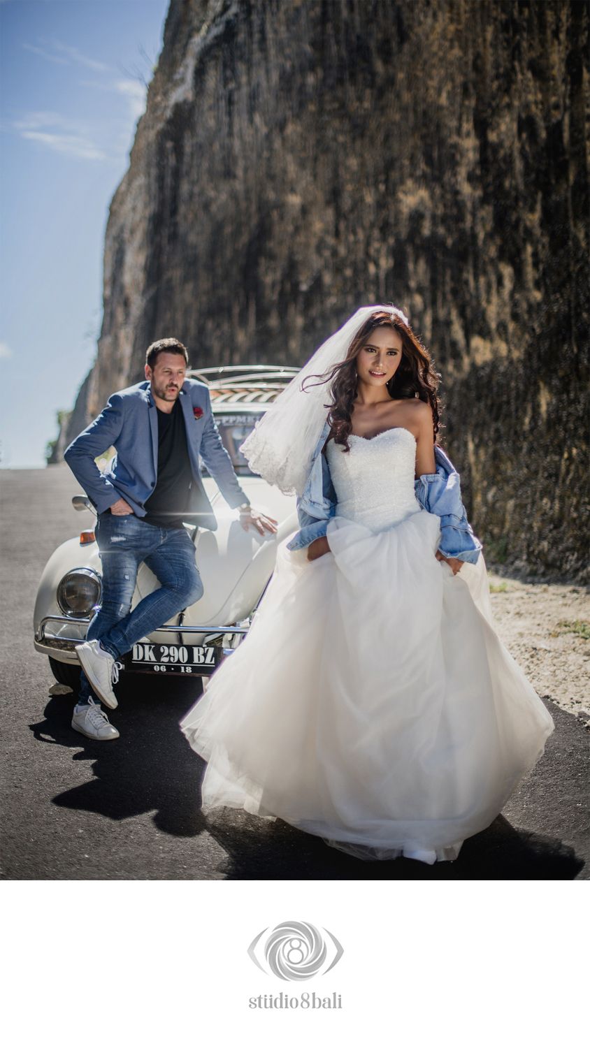 Bride in white gown with denim jacket and groom in blue suit pose by vintage car near cliffs in Bali.