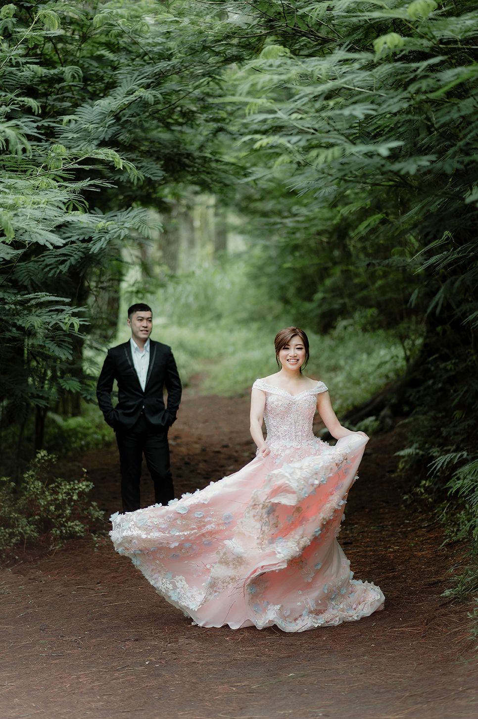Bride twirling in a pink lace gown on a forest path, groom in black suit standing behind her.
