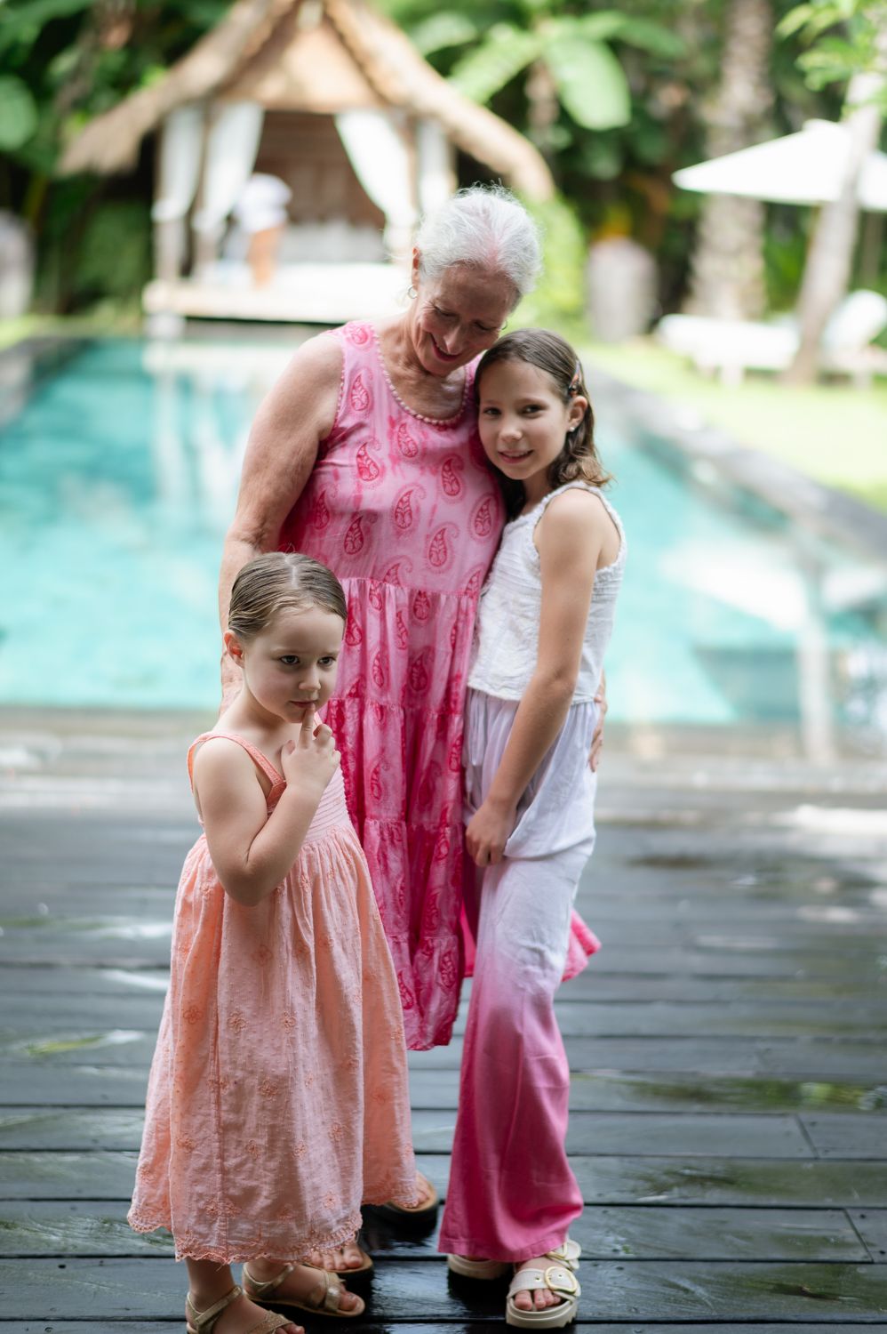 Grandmother in pink dress posing poolside with two young granddaughters in a tropical resort setting.