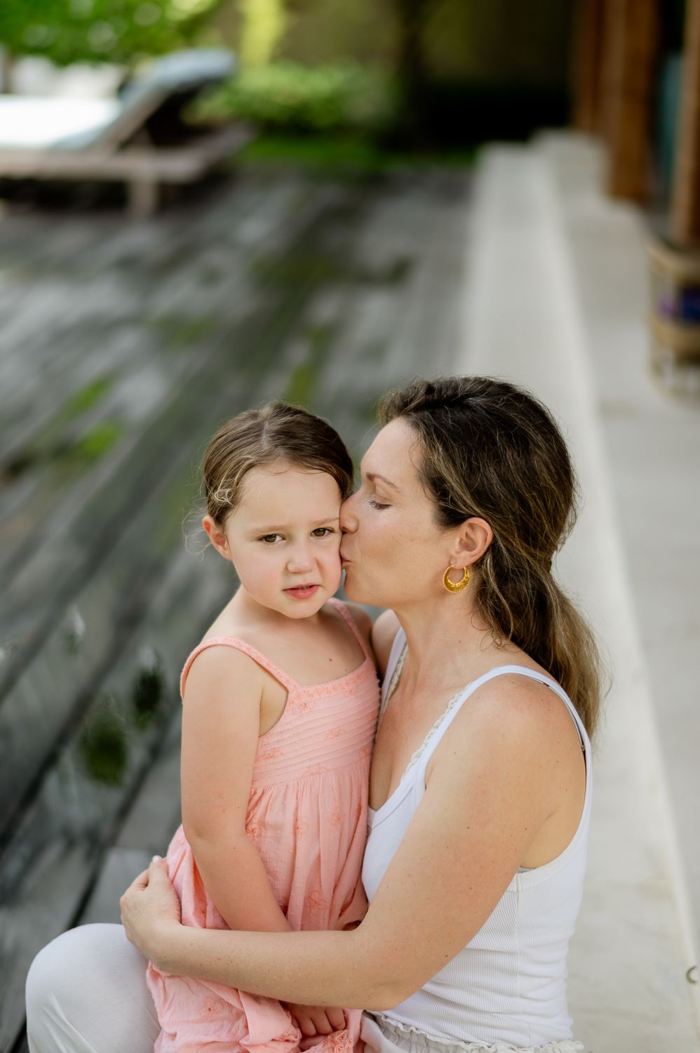 A mother tenderly kisses her young daughter's cheek while embracing her outdoors.