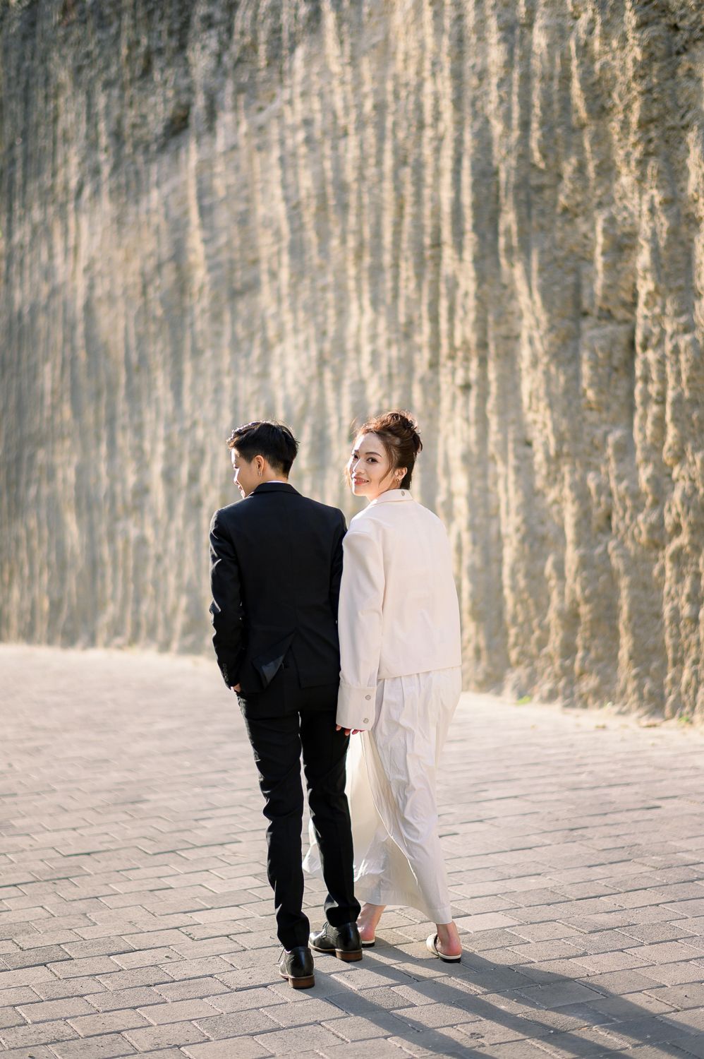 A couple in formal attire walks along a stone pathway, the woman in white glancing back while the man in black faces away.