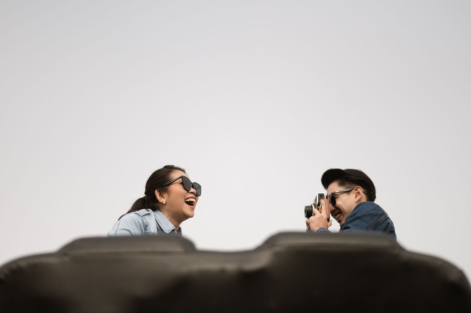 Two people laughing and taking photos outdoors against a gray sky, one wearing sunglasses and one holding a camera.