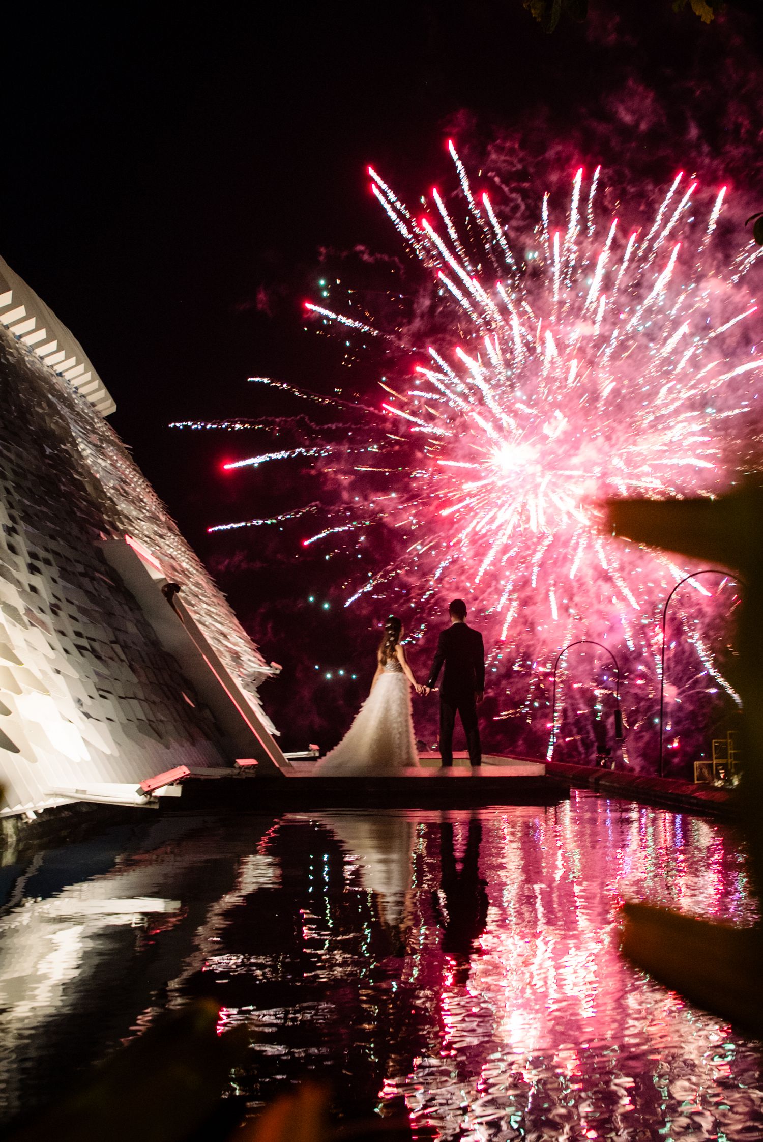 Bride and groom stand hand in hand on a waterside platform watching pink fireworks light up the night sky, reflected in the water.