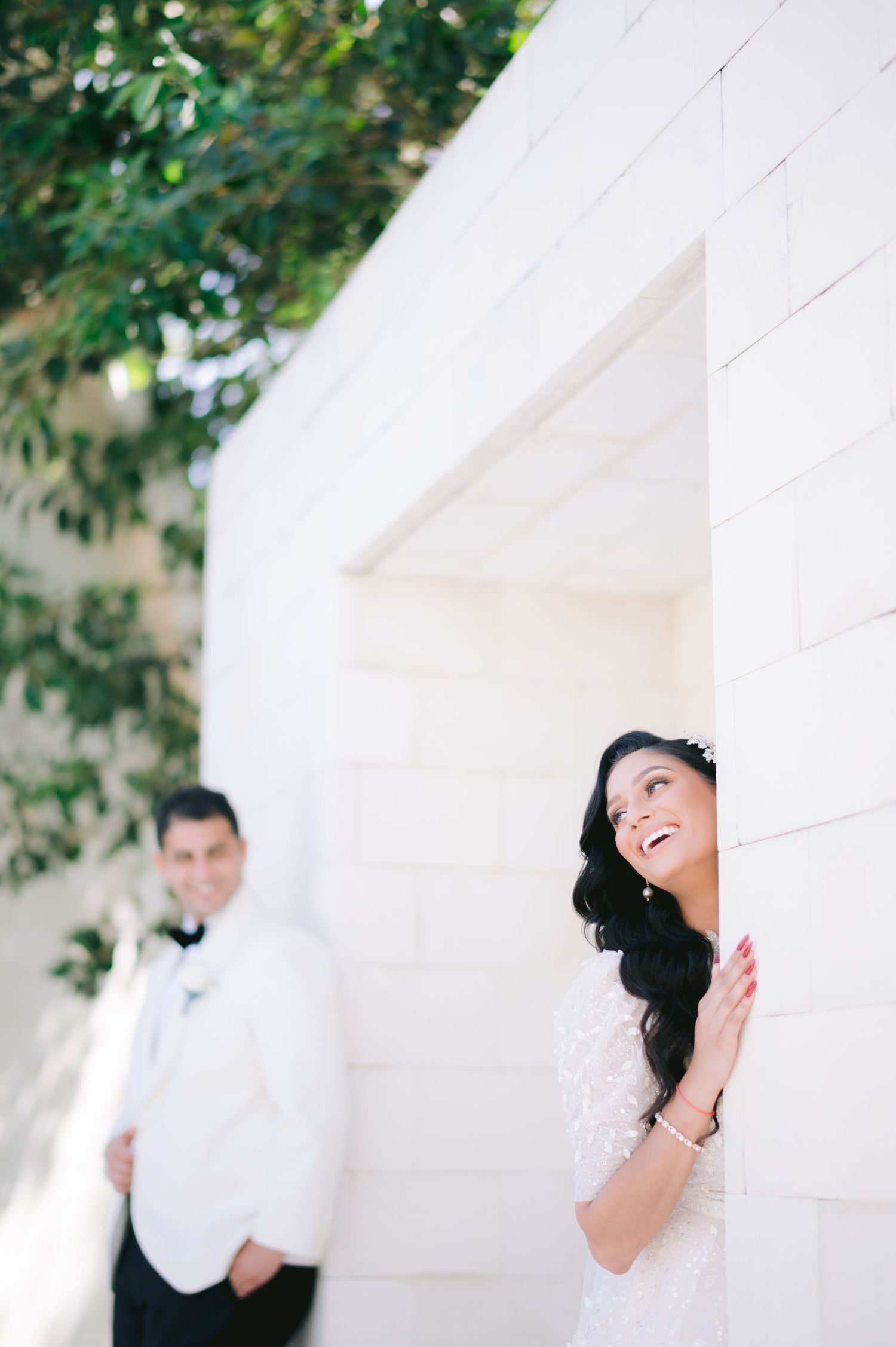 Bride in lace gown laughing by white wall while groom in white tuxedo stands blurred in background.