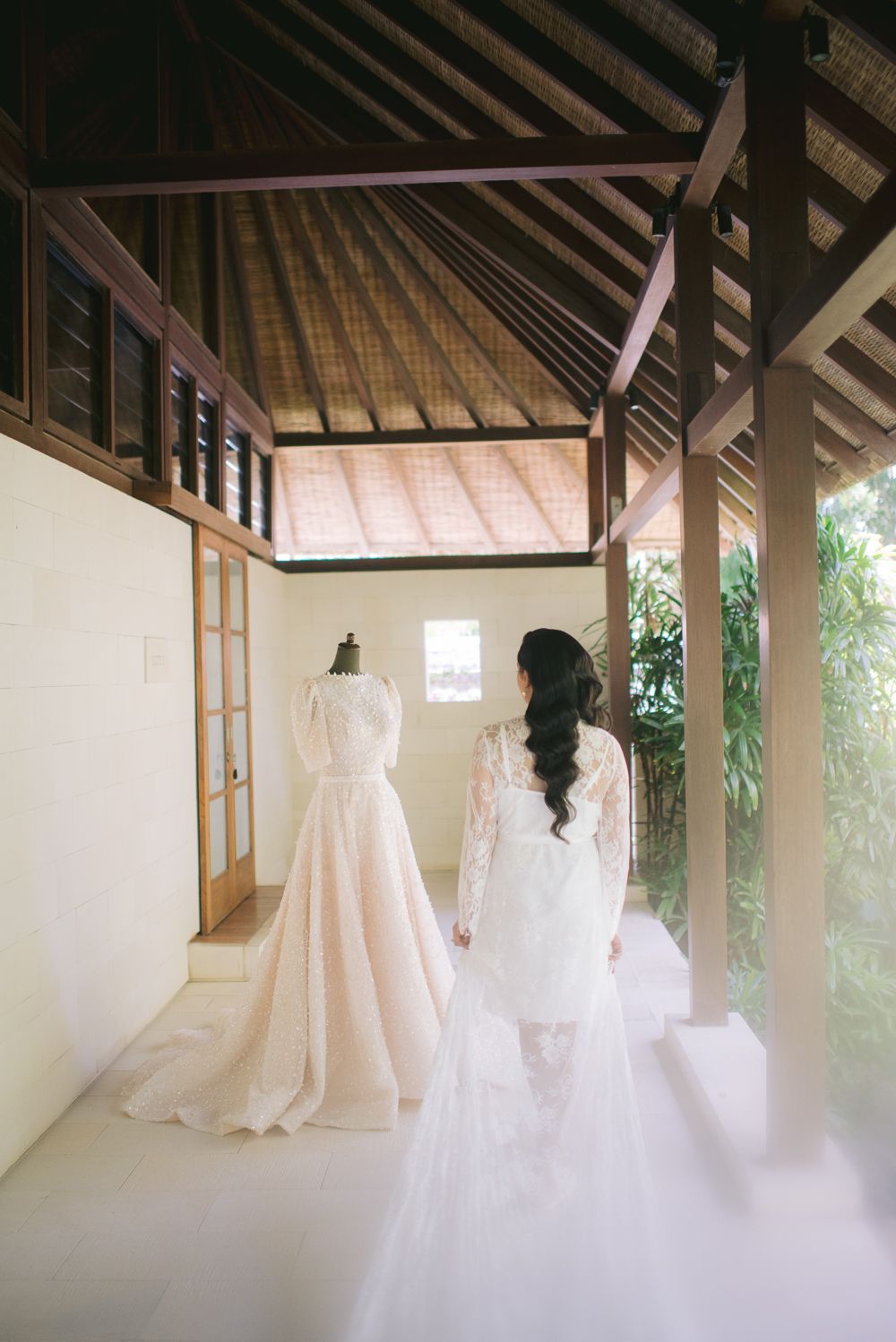 Bride in lace wedding gown views dress on mannequin in elegant Balinese open-air pavilion with wooden architecture.