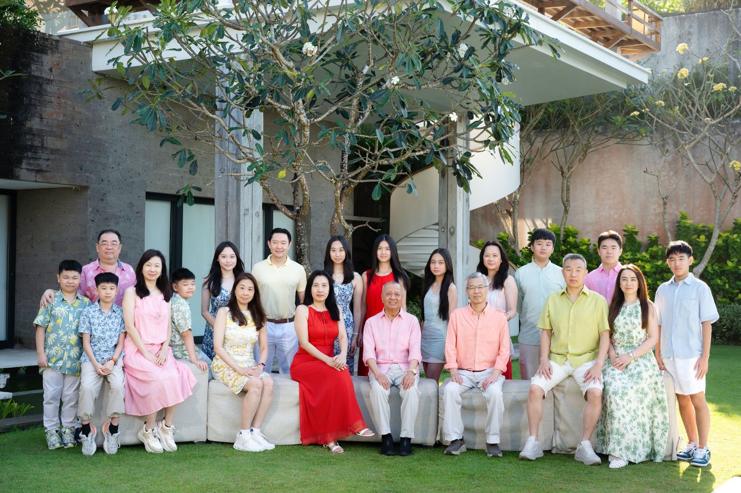Large Asian family group portrait outdoors in garden, multi-generational gathering with adults and children, lush greenery background.