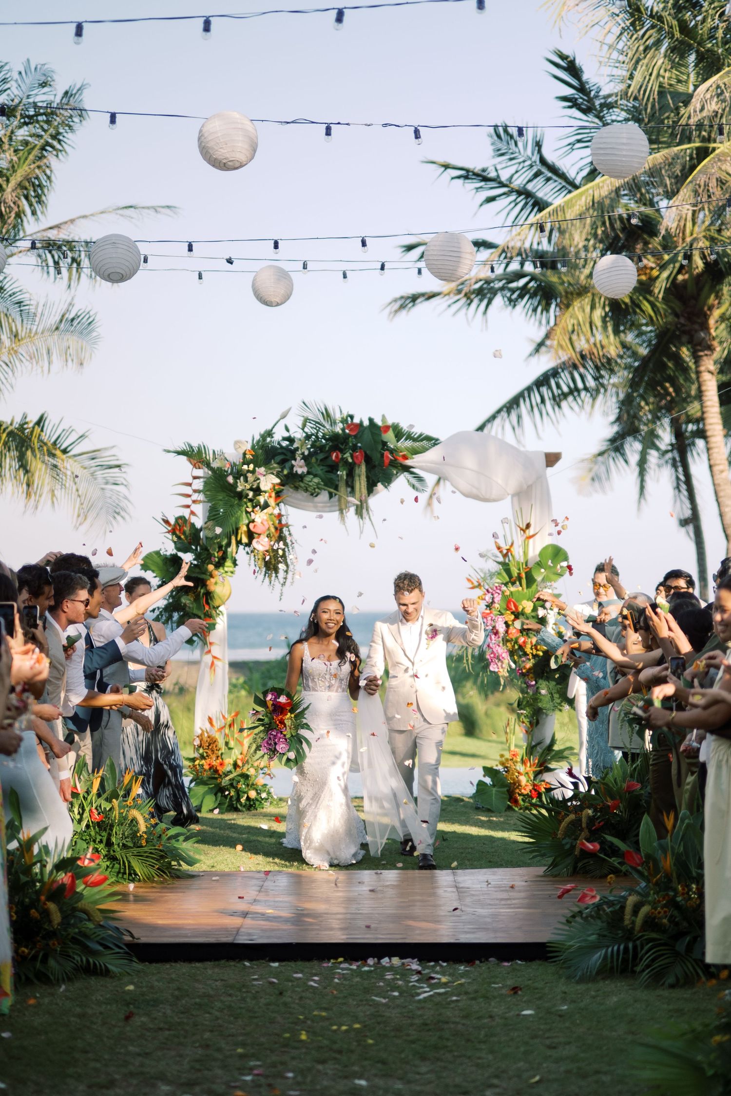 Newlywed couple walks down tropical outdoor aisle under paper lanterns, surrounded by lush floral arch and cheering guests.