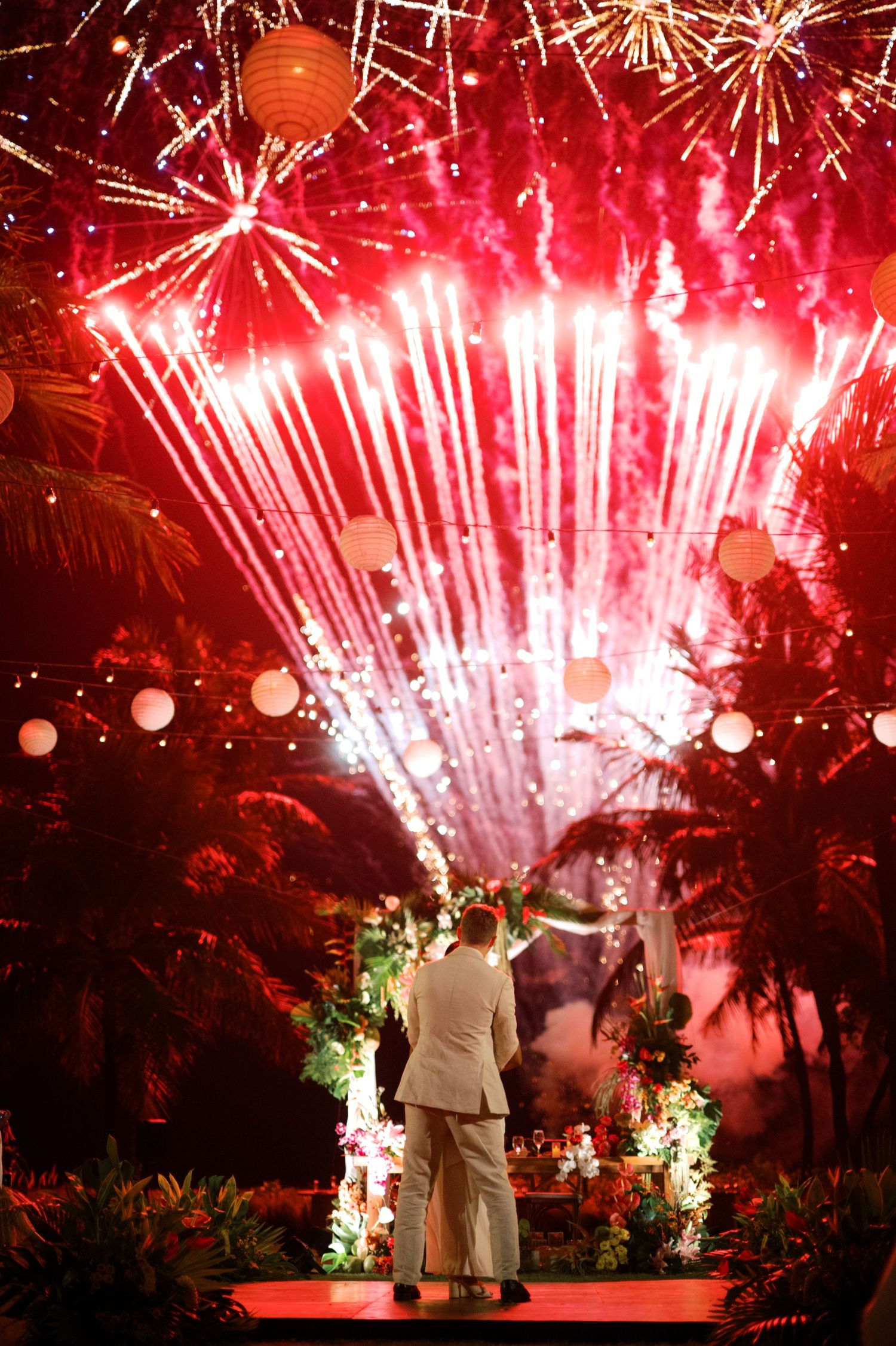 Couple stands before stunning red fireworks display at tropical outdoor wedding celebration at night.