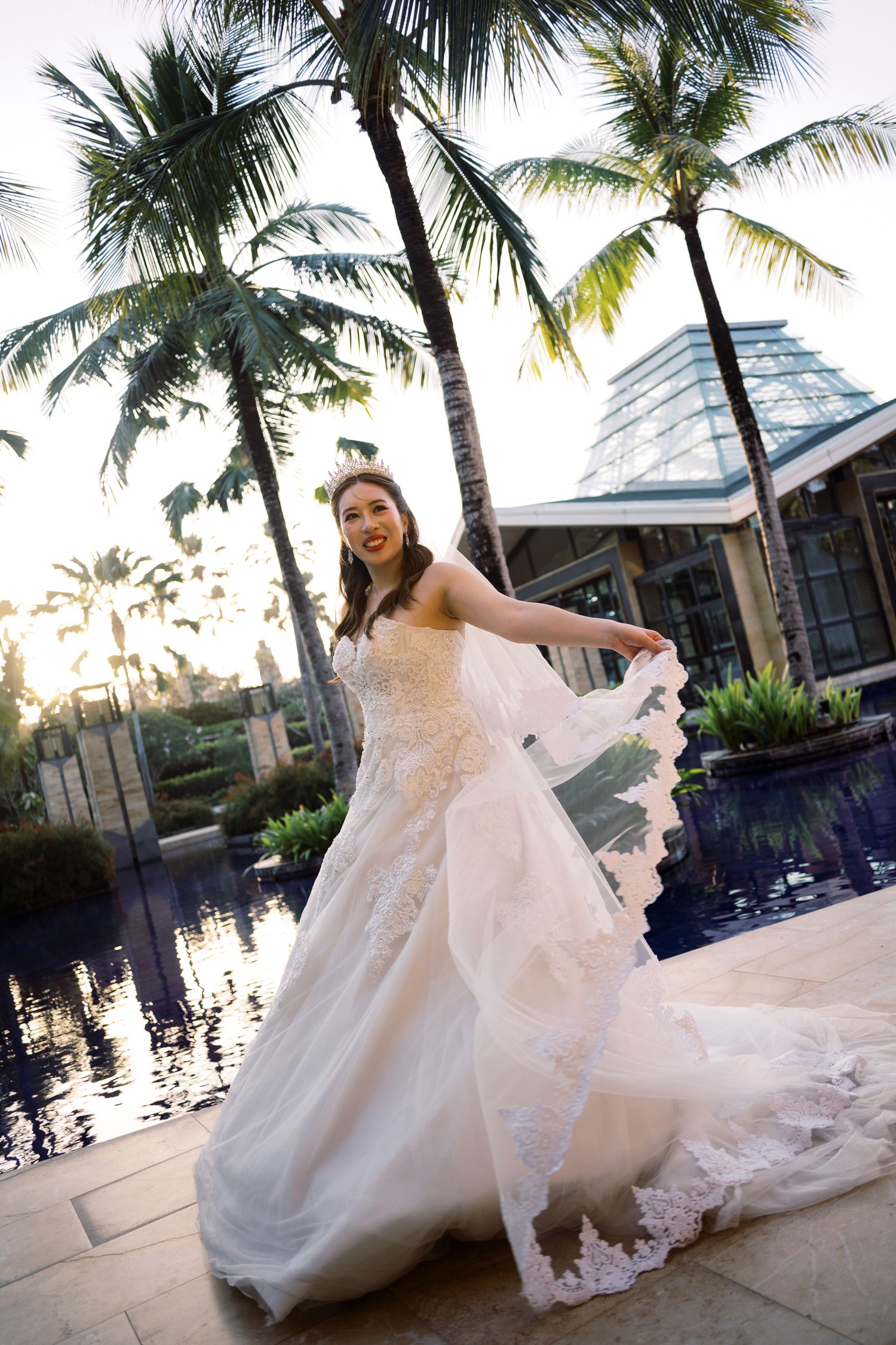 Bride in lace wedding gown twirling near pool with palm trees at tropical resort venue at sunset.