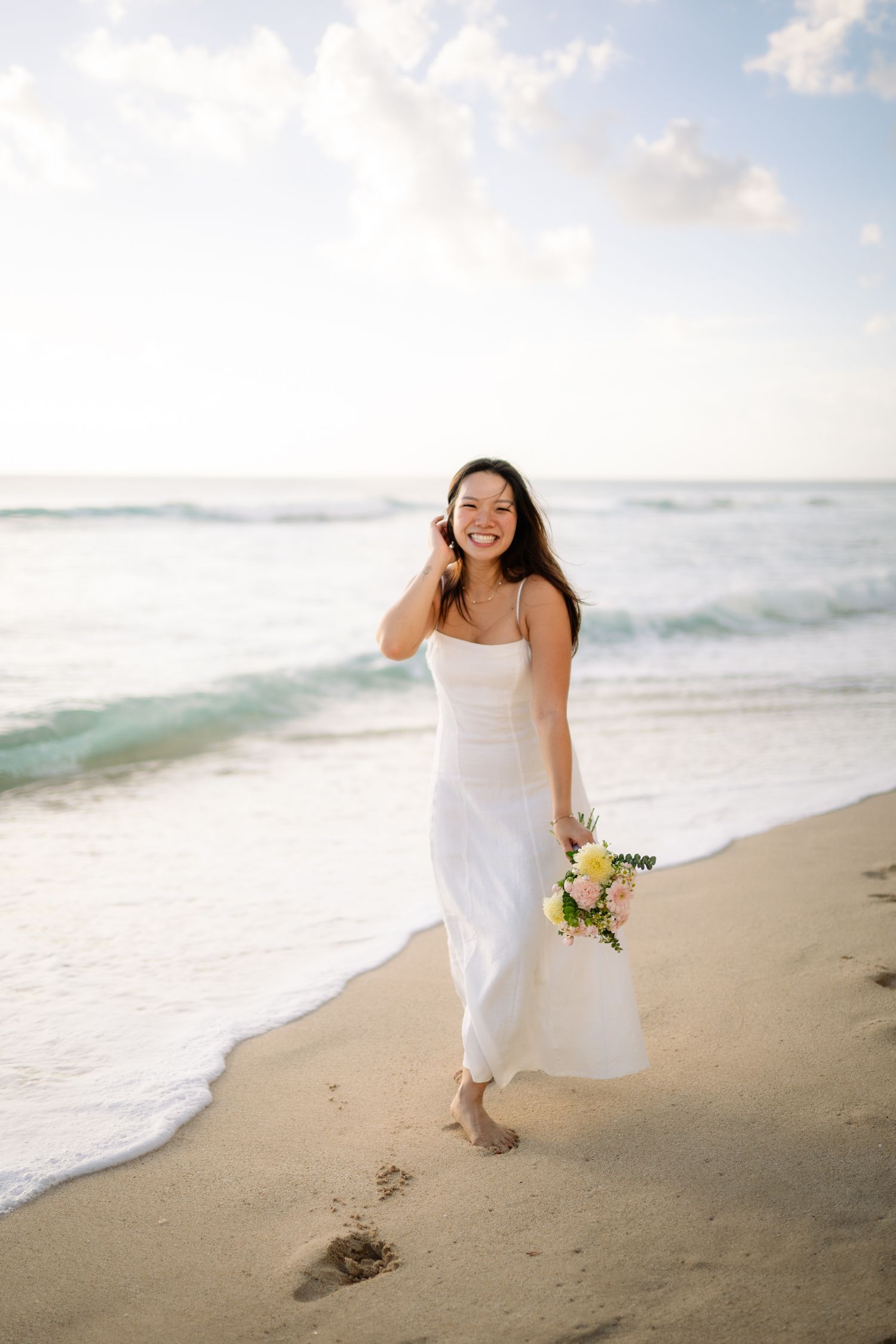 A smiling bride in a white dress walks barefoot on a sandy beach holding a colorful bouquet with ocean waves behind her.