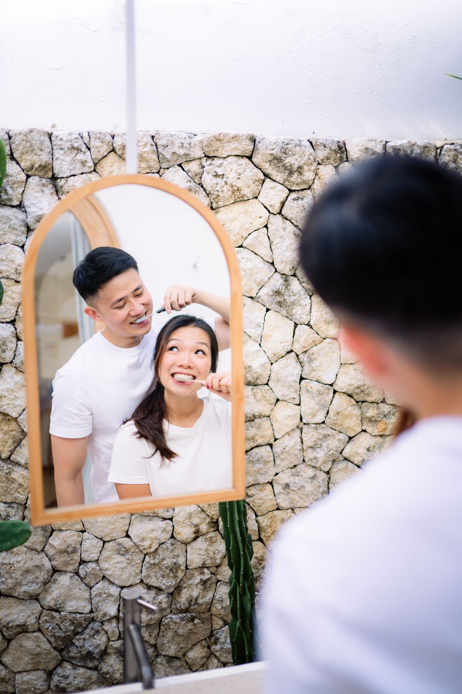 Asian couple in white shirts brushing teeth together, smiling at bathroom mirror with stone wall backdrop.