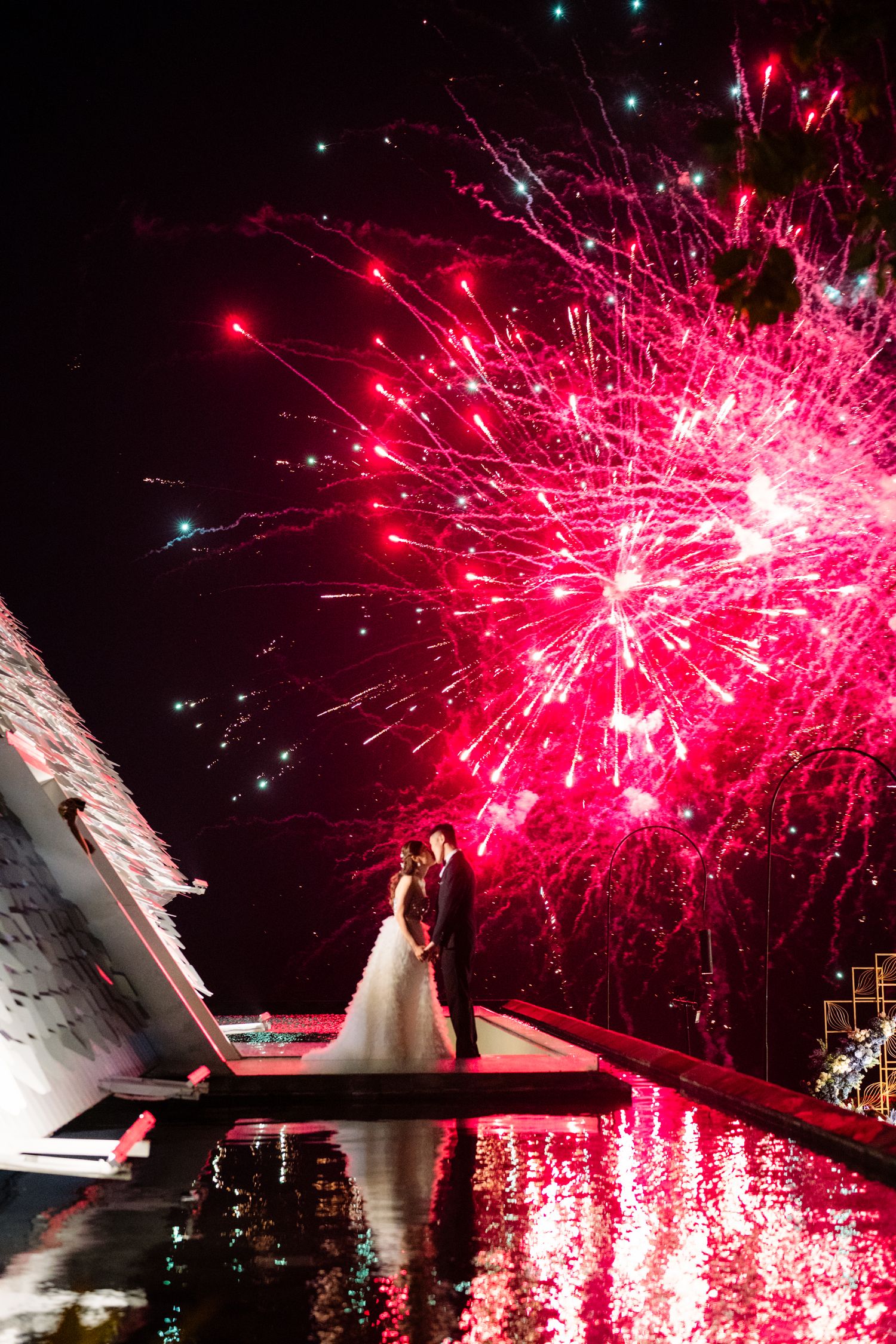 Bride and groom embrace on a waterside platform at night as vivid pink fireworks burst overhead and reflect in the water.