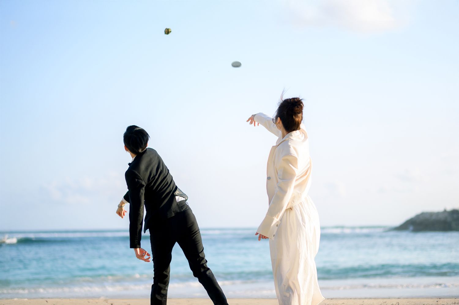 Couple in wedding attire playing on a sandy beach, tossing a ball with ocean waves in the background.