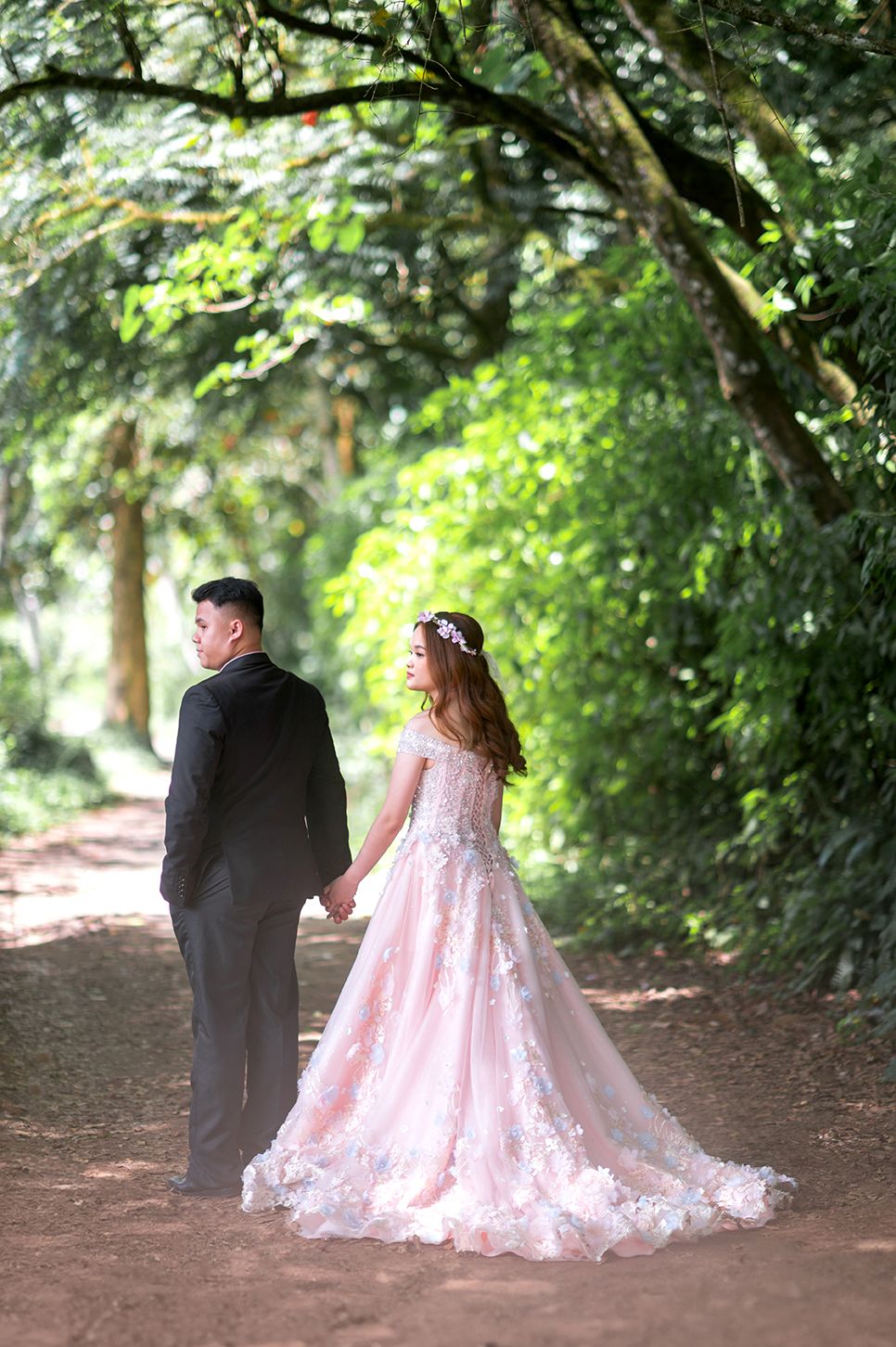 A couple in wedding attire holds hands on a tree-lined path, bride in pink floral gown with floral crown.