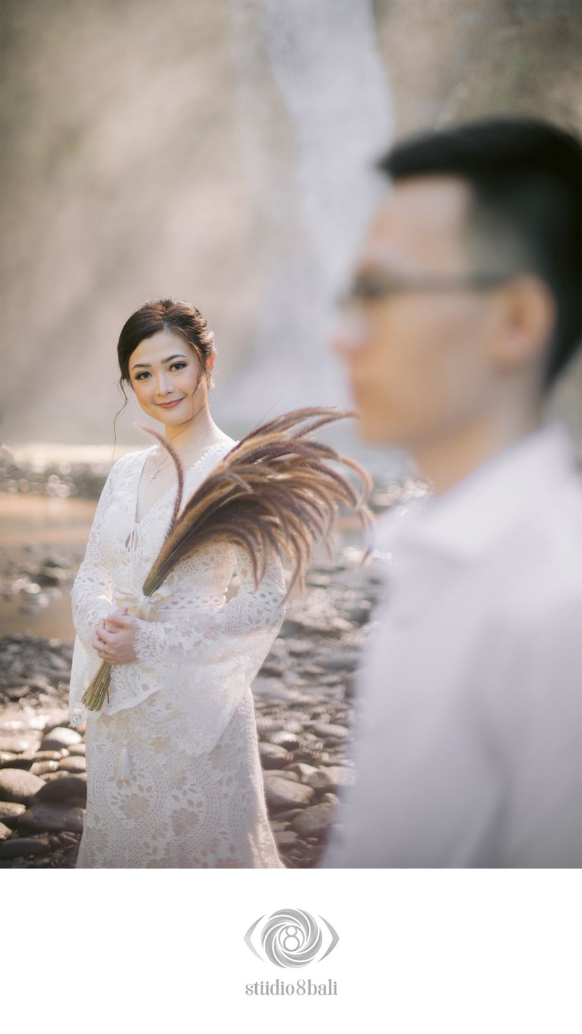 Bride in white lace dress holds dried floral bouquet near waterfall, groom blurred in foreground, romantic pre-wedding shoot.