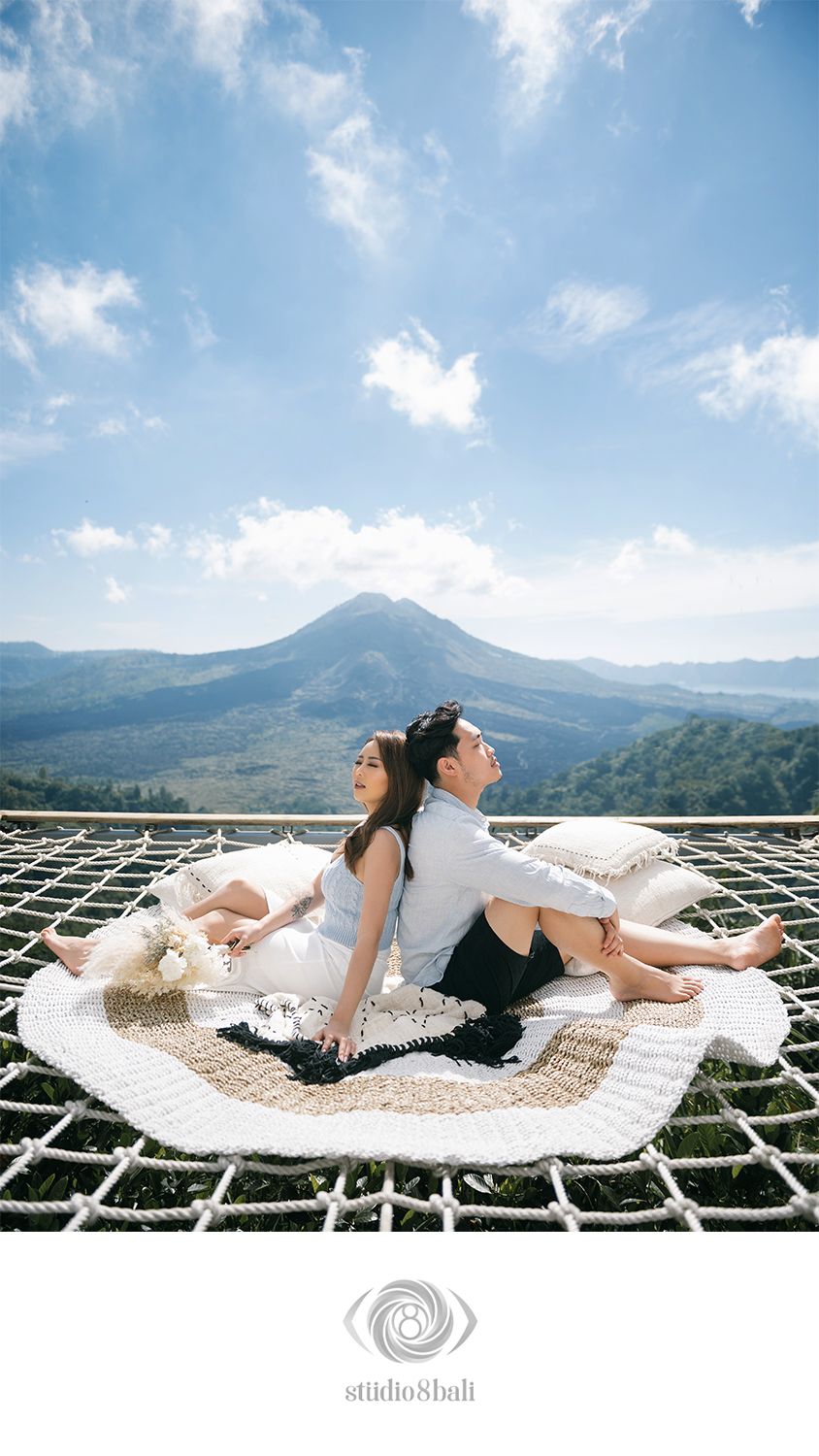 Couple sitting back-to-back on a rope net with Mount Batur volcano backdrop in Bali, Indonesia.