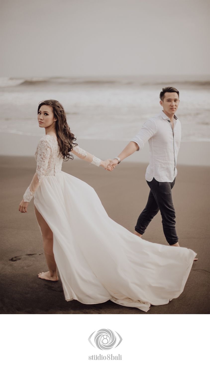 Couple walking on dark beach, bride in flowing white lace gown holding groom's hand, ocean waves in background.