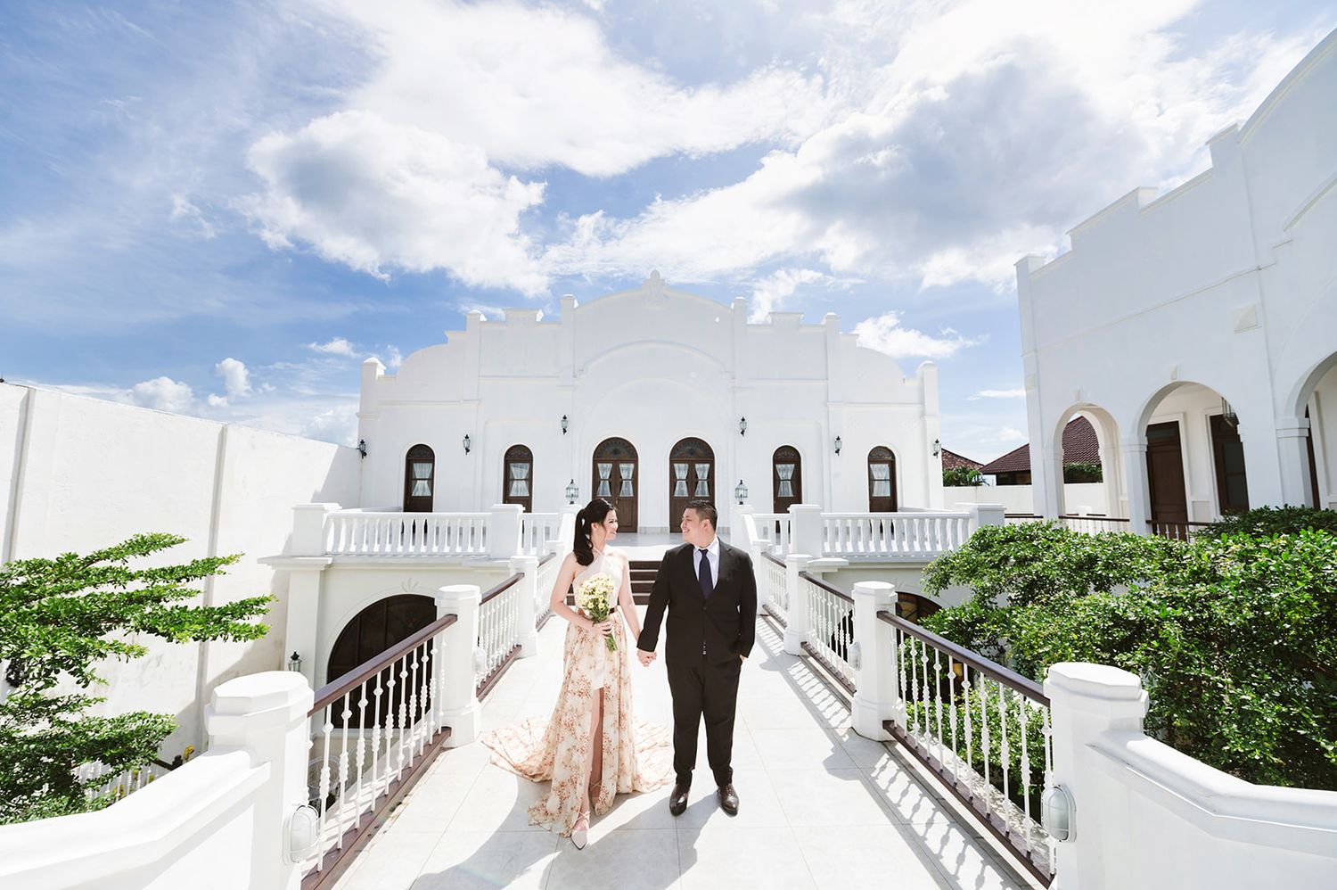 Bride and groom walk hand in hand on a white balcony terrace of an elegant wedding venue under blue skies.
