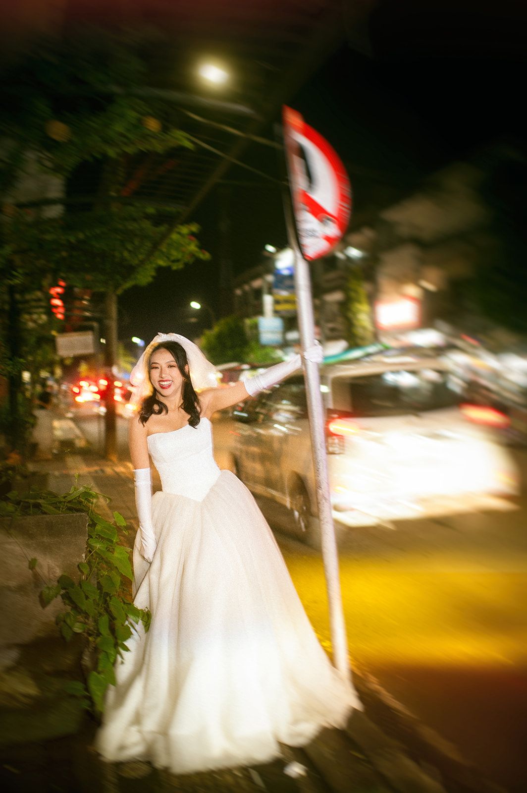 Smiling bride in white gown posing beside a street sign on a busy urban road at night.