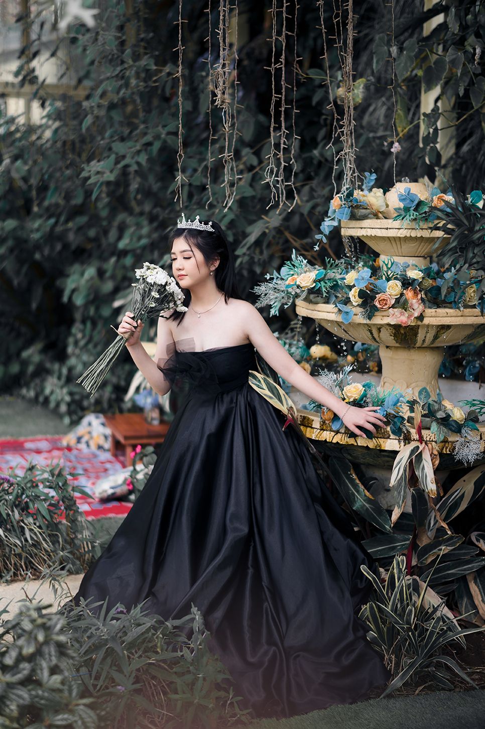 Woman in black strapless ball gown and crown holds white bouquet beside floral fountain in lush garden.