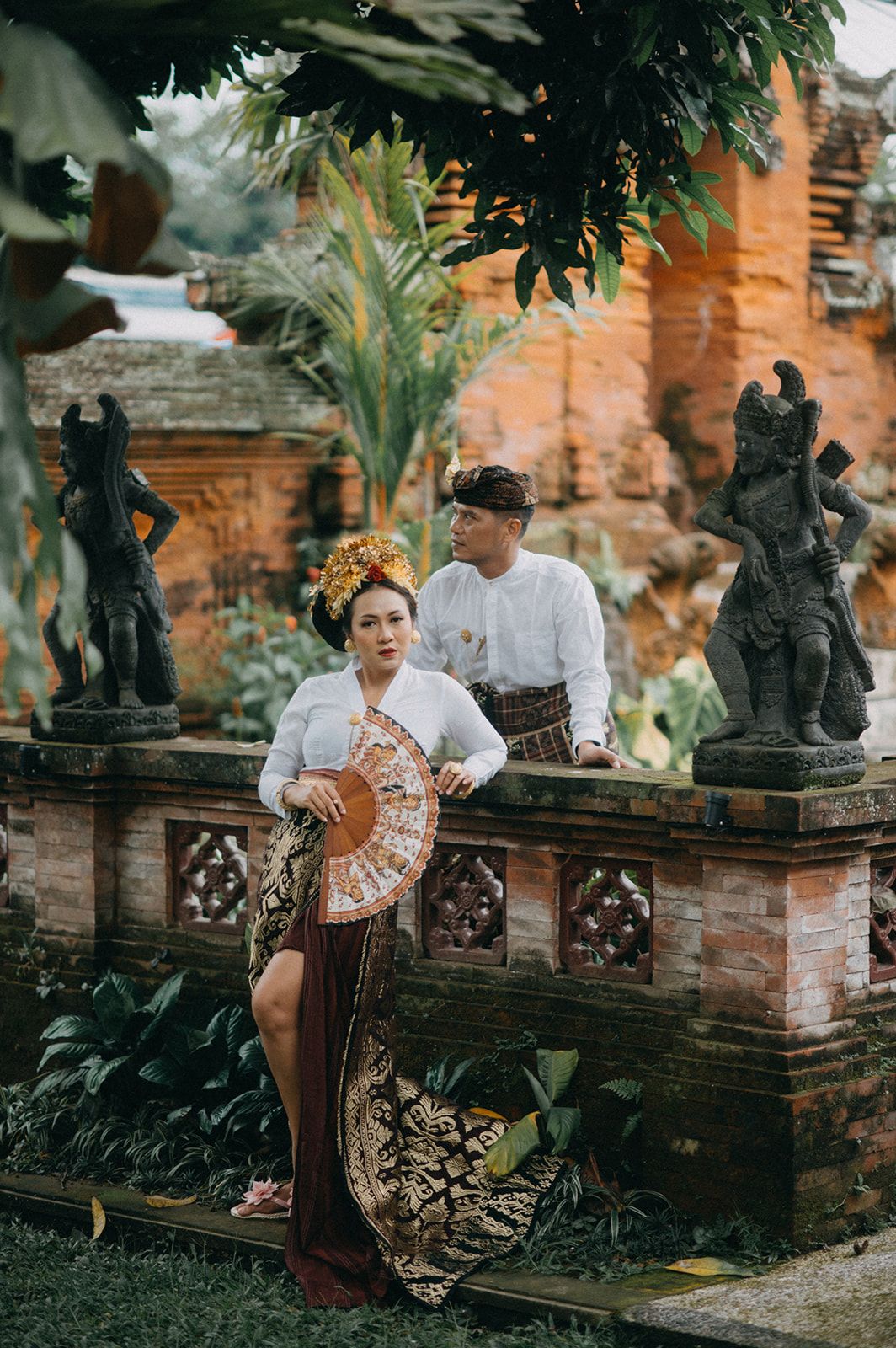 Balinese couple in traditional attire posing at ornate temple with stone statues and lush greenery.