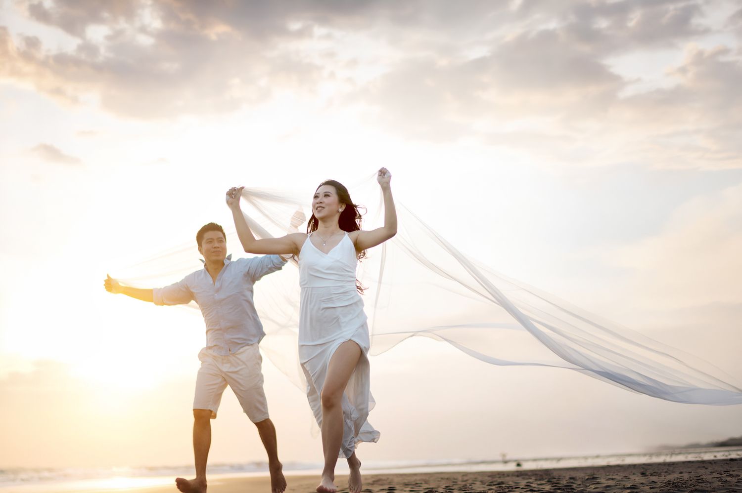 Happy couple in white running on a beach at sunset, bride's veil flowing in the wind.