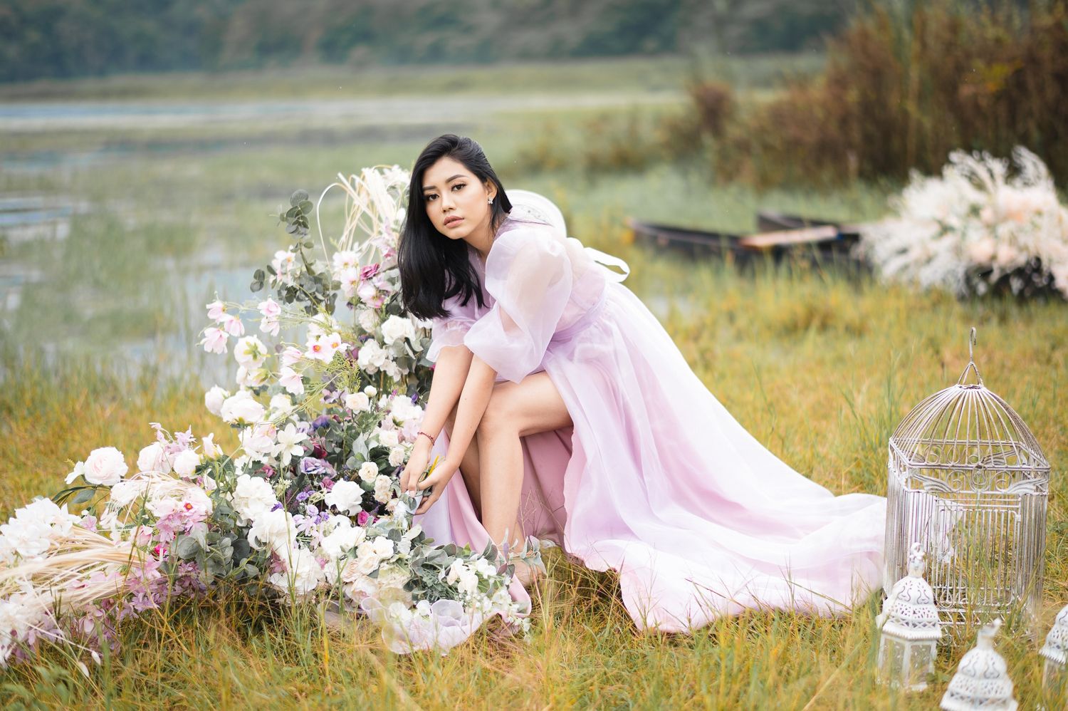 Woman in pink sheer gown poses among floral arrangements in a serene outdoor setting near water.
