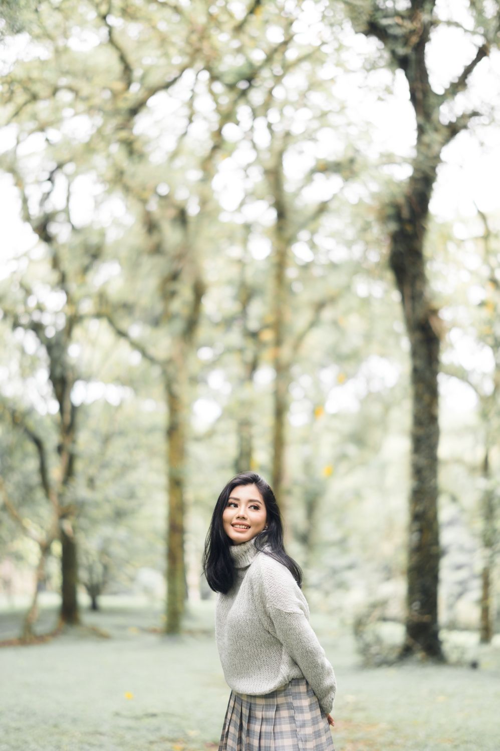 Young Asian woman in gray turtleneck sweater and plaid skirt smiling in a serene tree-lined park.
