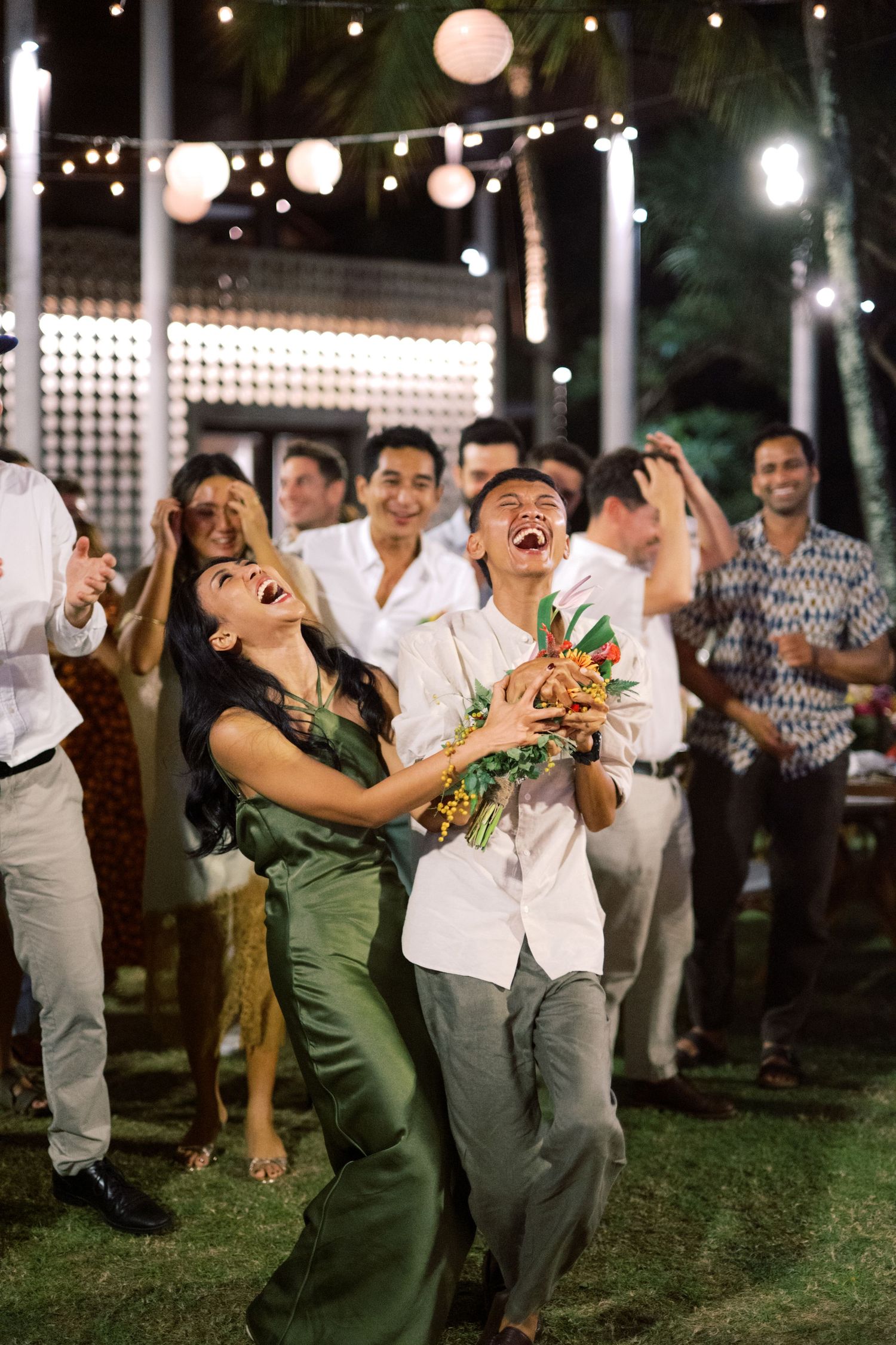 Excited couple catches bridal bouquet together at tropical outdoor wedding reception surrounded by laughing guests.