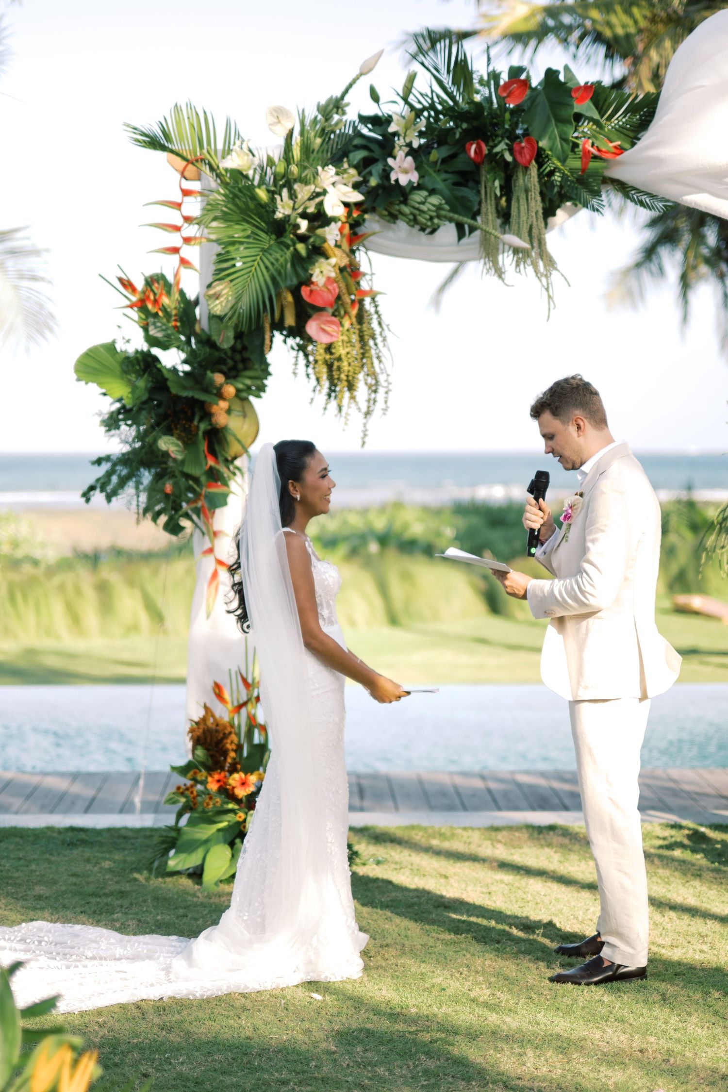 Bride and groom exchange vows under tropical floral arch with ocean view at outdoor ceremony.