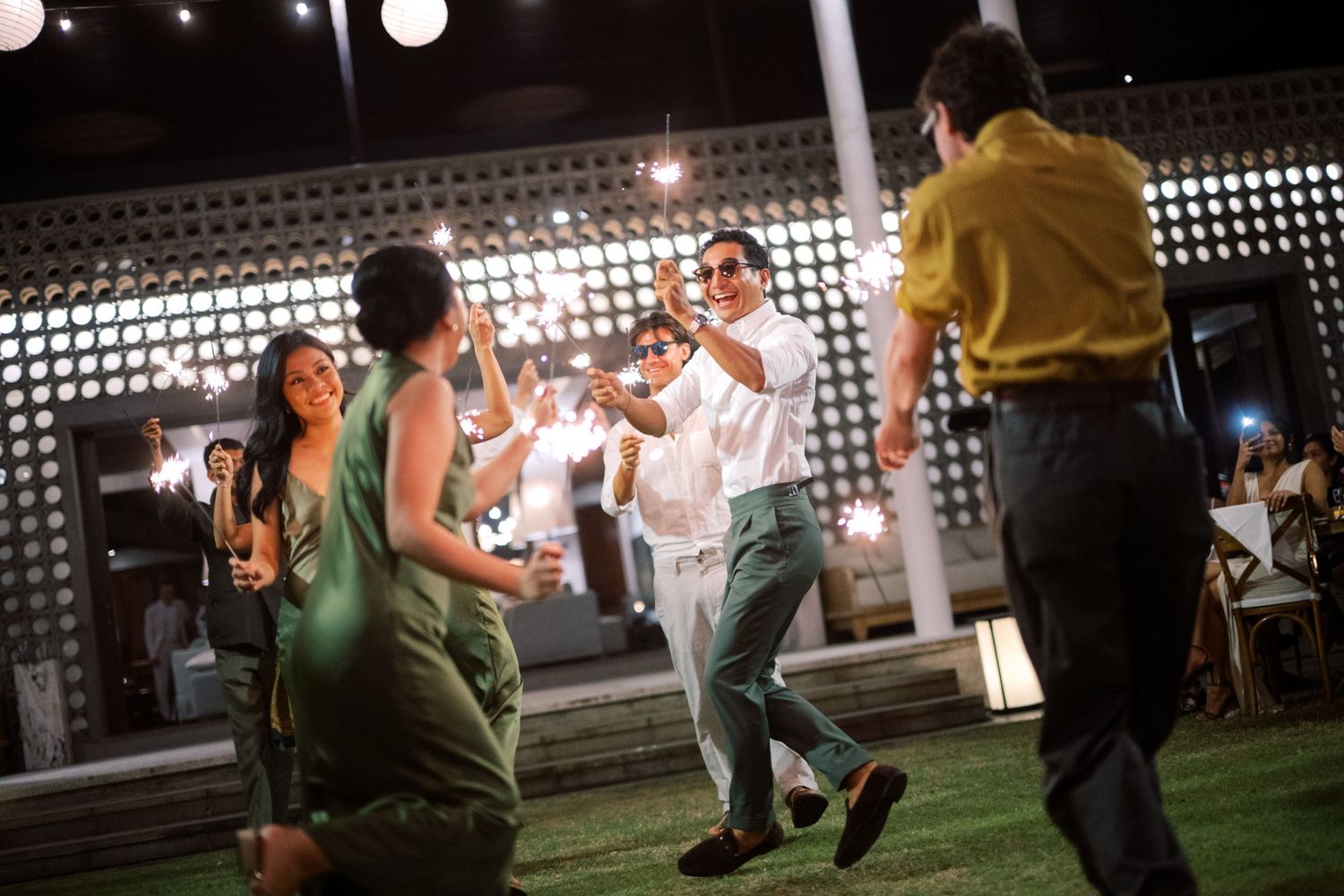 Guests dance energetically under string lights at an outdoor tropical wedding reception at night.