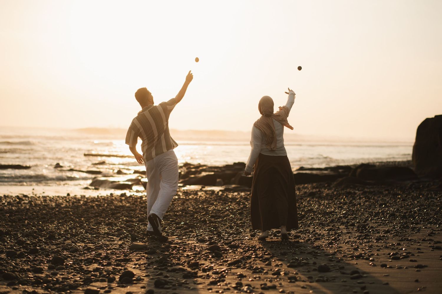 Two people joyfully tossing rocks on a rocky beach at sunset with ocean waves in the background.