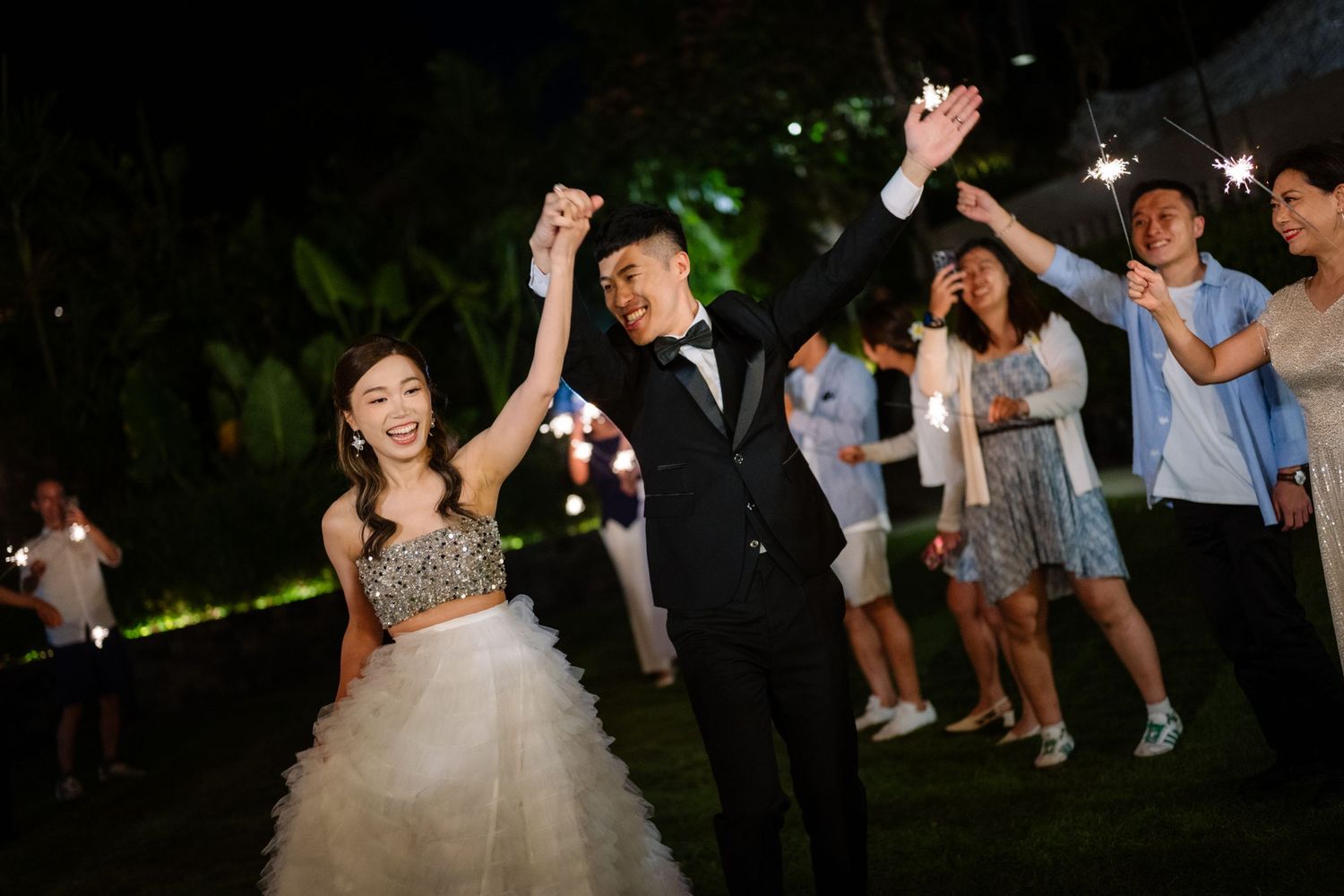 Bride in a sparkling gown and groom in a black suit hold hands while guests cheer with sparklers at an evening outdoor wedding.